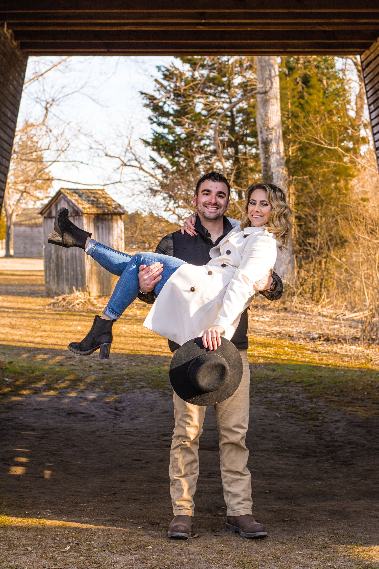 Man carrying woman in white coat outdoors at rustic farm setting during golden hour, holding a hat.
