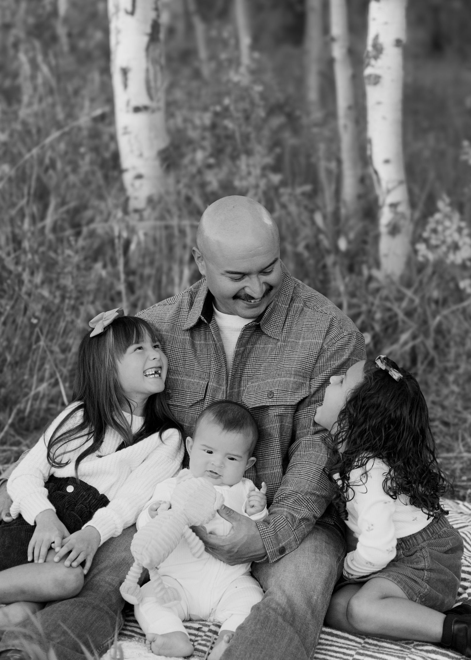 Black and white photo of a bald father sitting outdoors with three young children among birch trees.