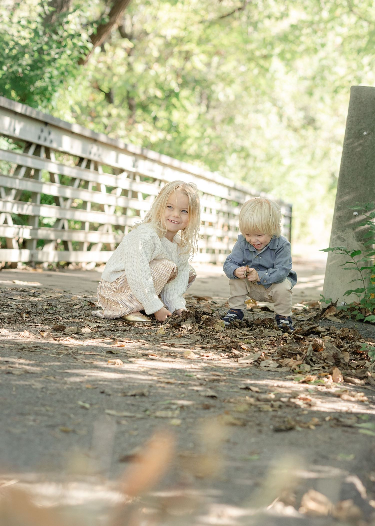 Two young blonde siblings crouch on a shaded path near a wooden bridge, playing with leaves during an outdoor photo session.