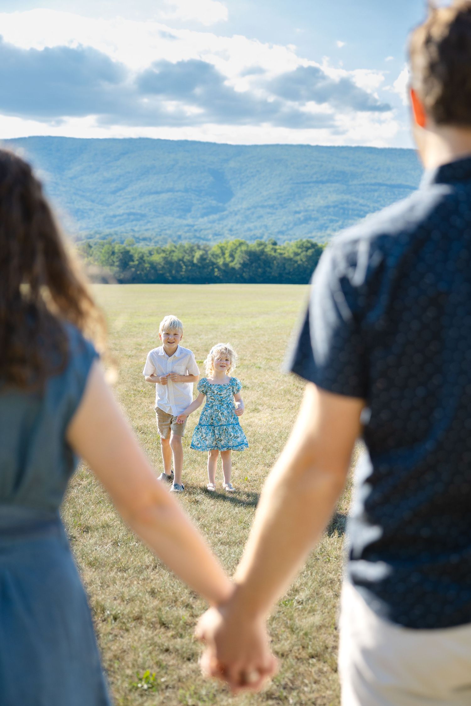 Parents holding hands watching two young children run toward them in a sunny green field with mountain views. Roanoke, VA