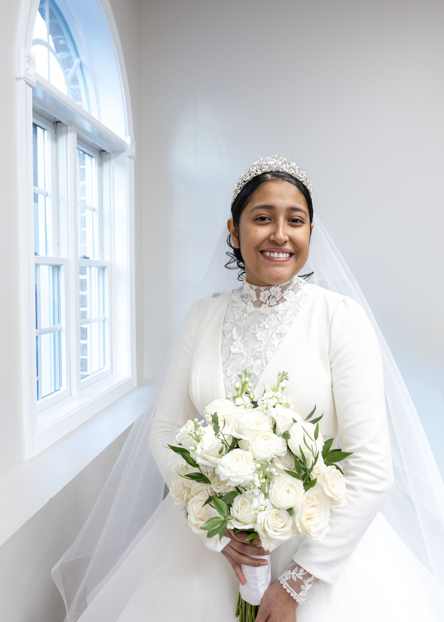Smiling bride in white lace gown, tiara, and veil holding white rose bouquet by a bright window.
