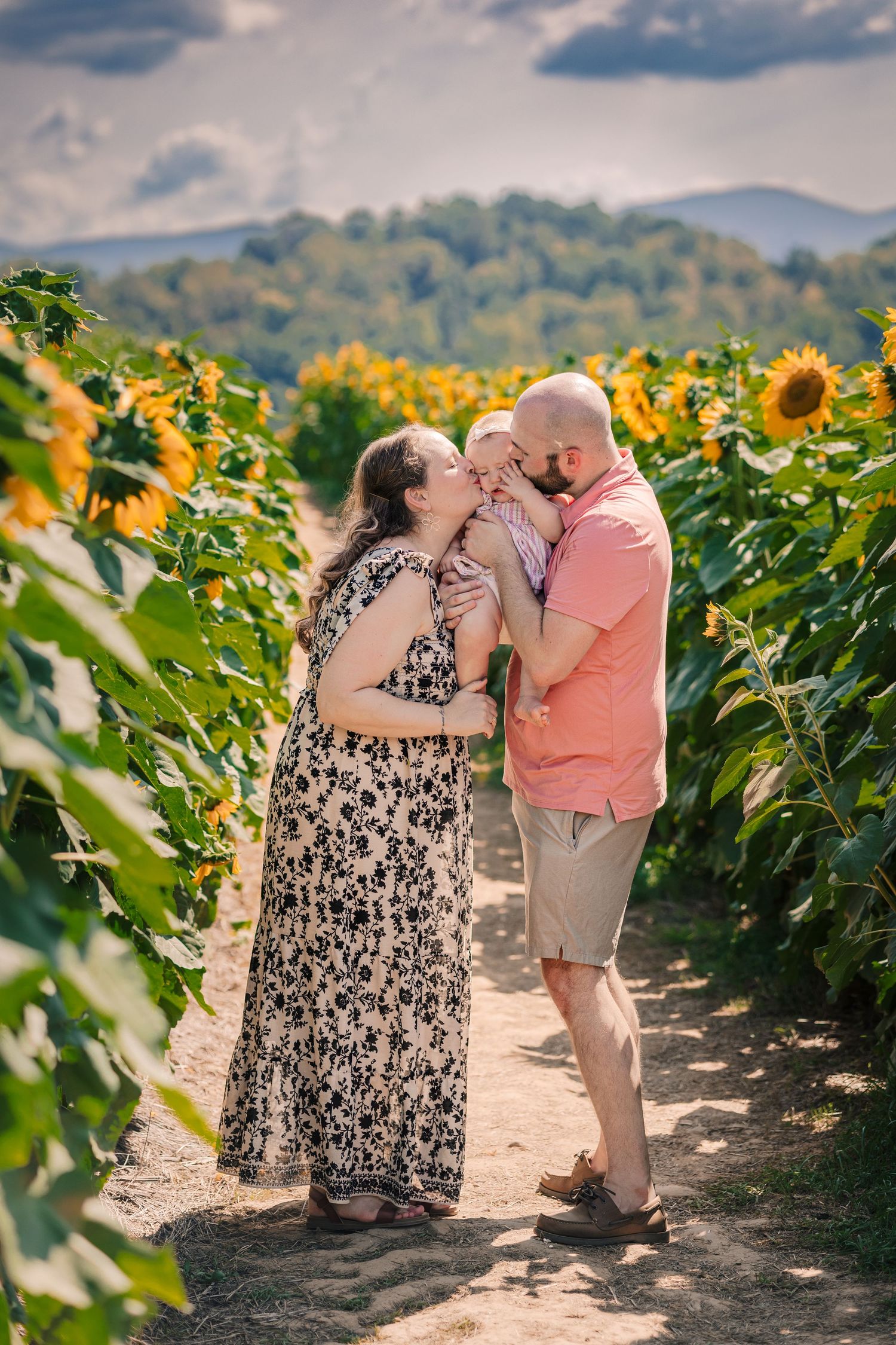 A couple shares a tender kiss with their baby in a sunflower field with mountain views.