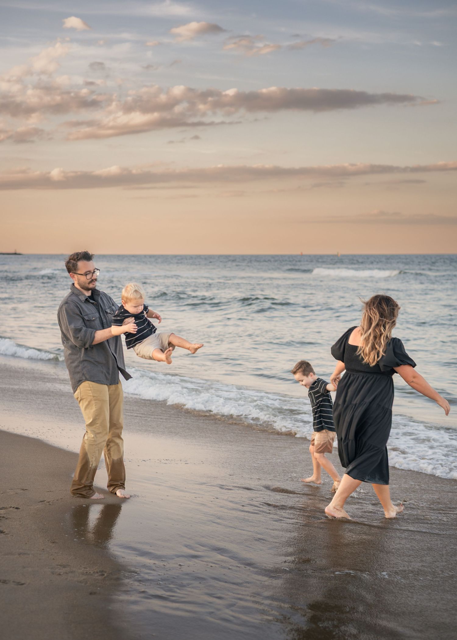 Family playing on beach at sunset, father lifting baby, mother holding toddler's hand by ocean waves.