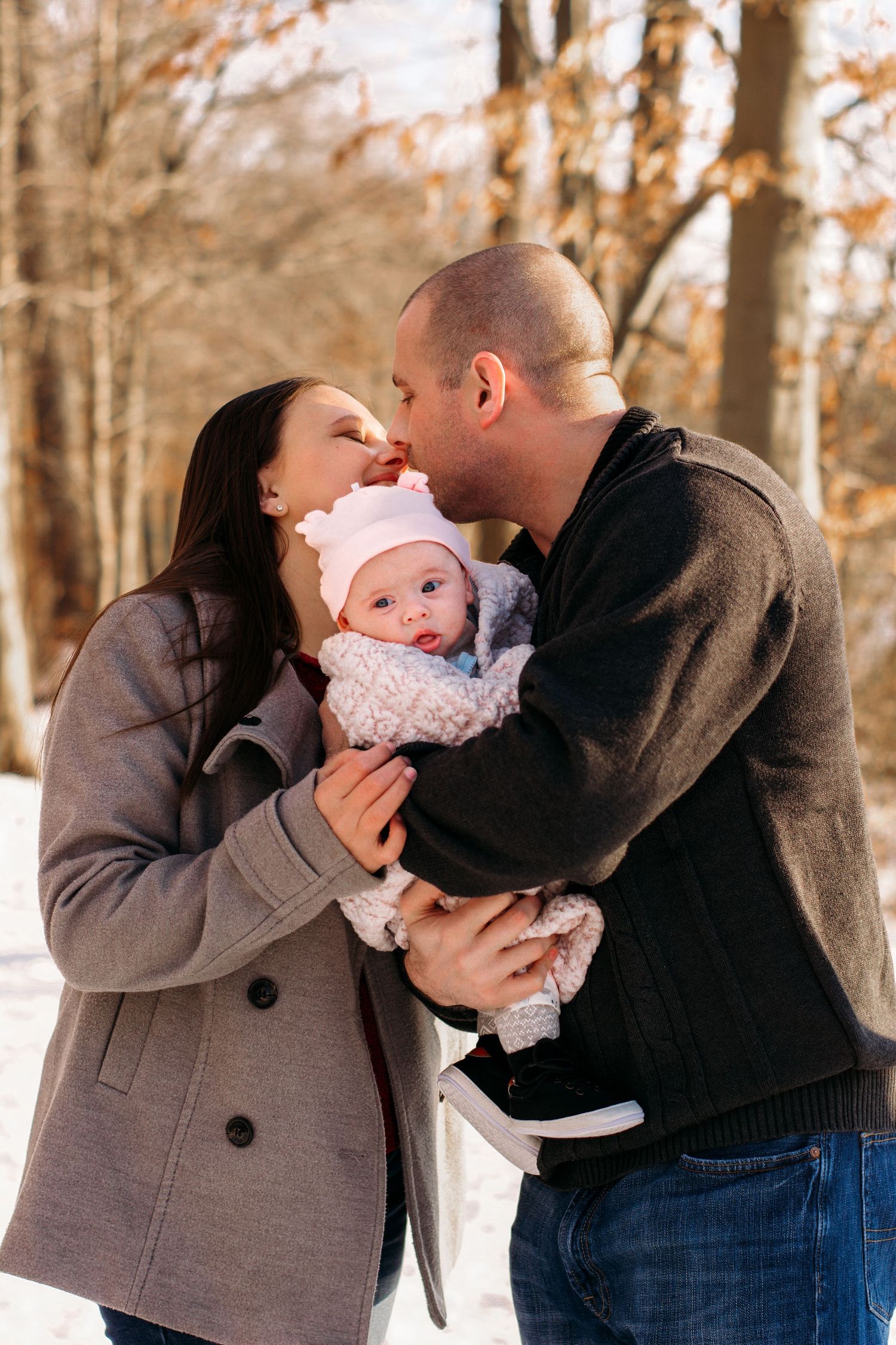 Parents kissing their baby in a snowy winter forest, family dressed in warm coats and pink hat outdoors.