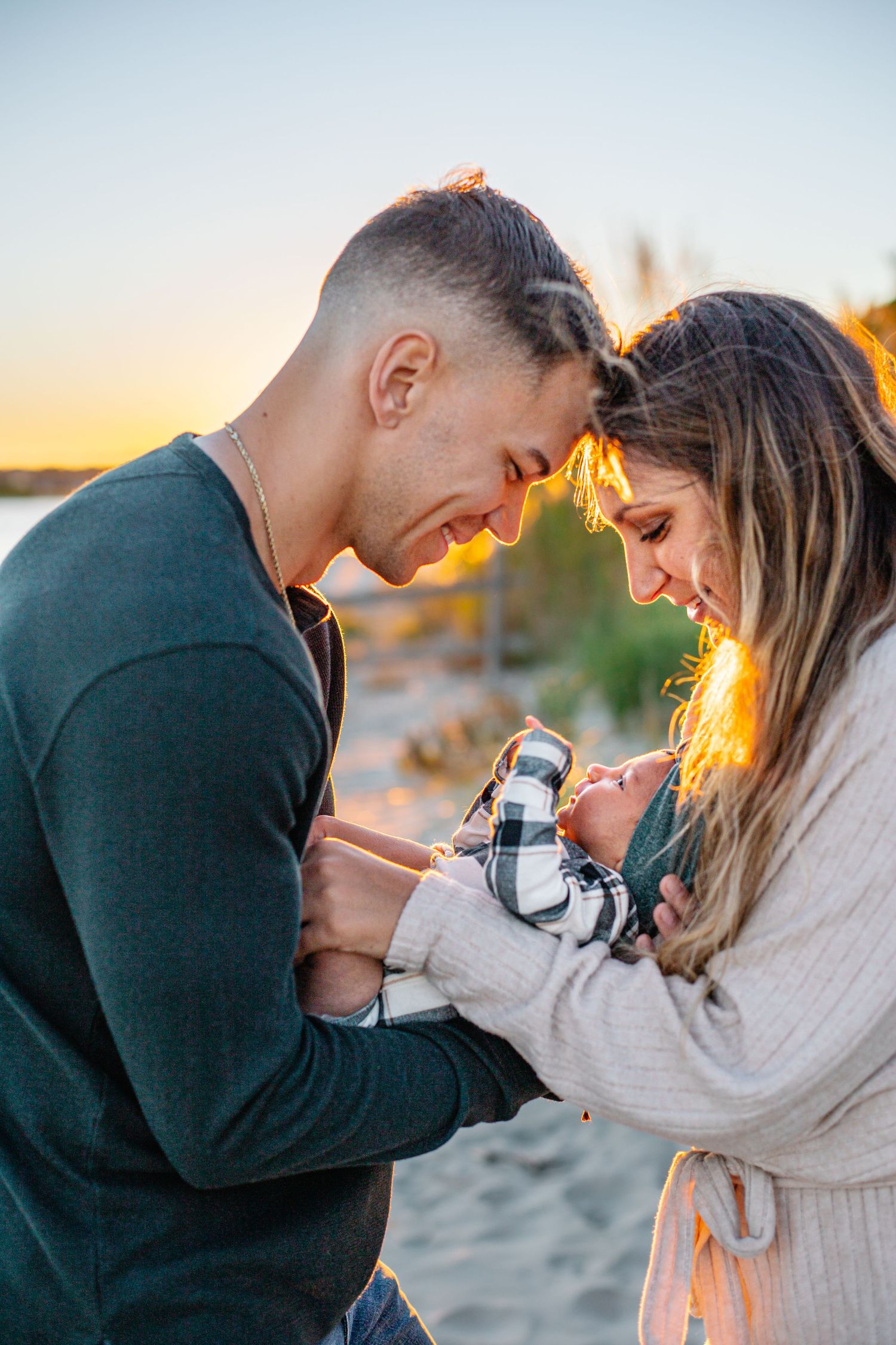 Couple touching foreheads and smiling at their newborn baby on a sandy beach during golden hour sunset.
