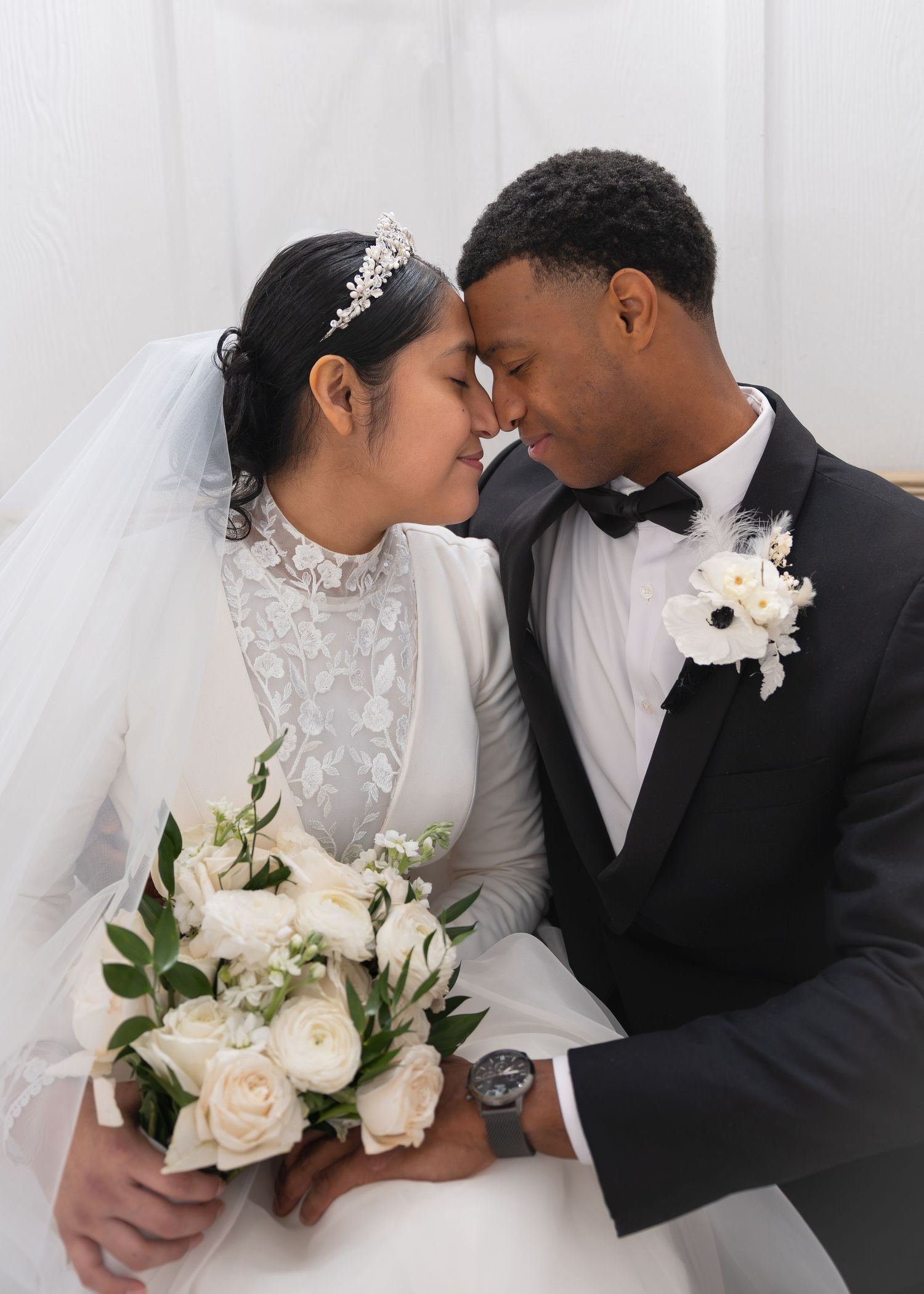 A bride and groom in wedding attire share an intimate moment, touching foreheads, with white roses bouquet.