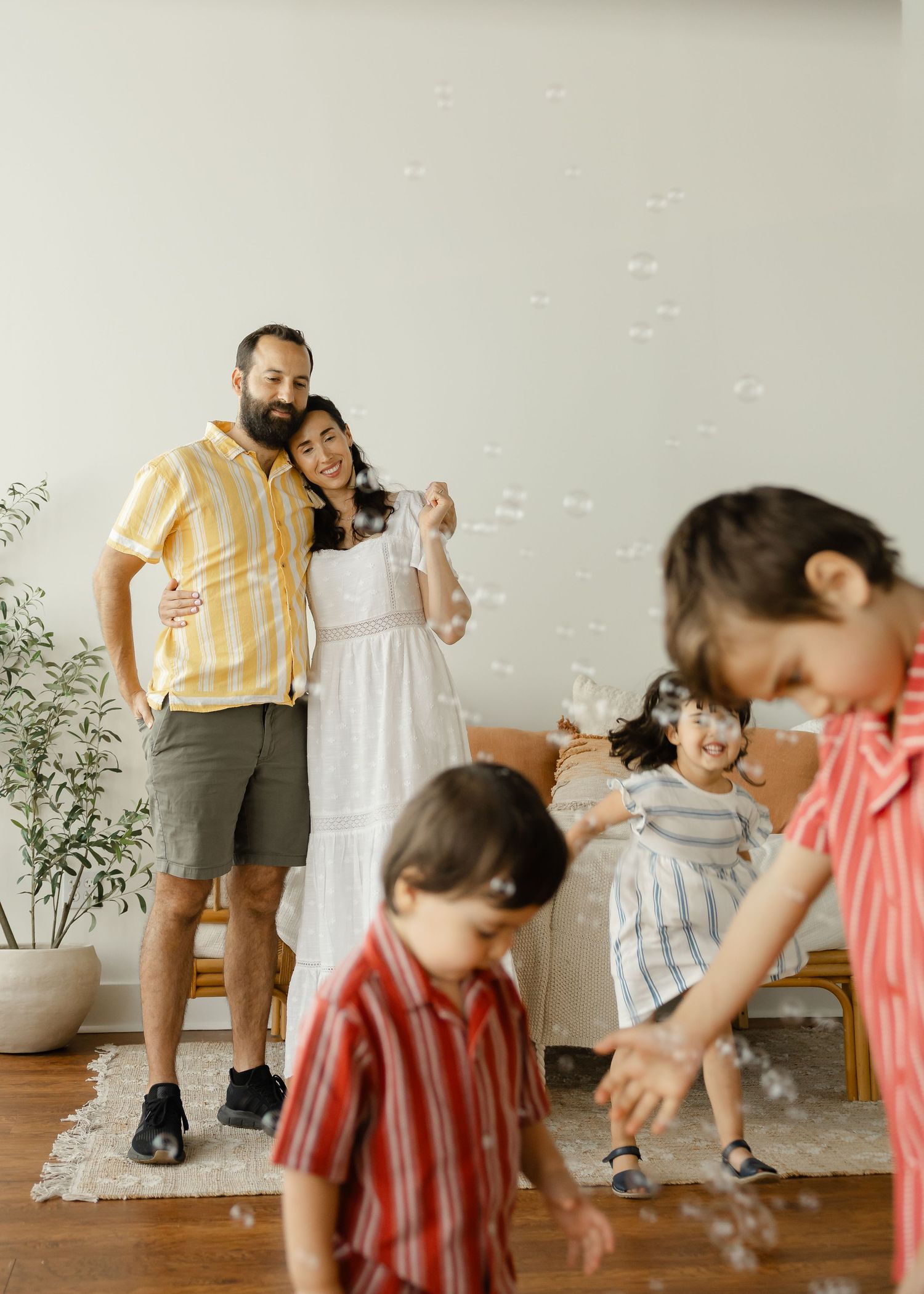 A bearded dad and mom in white dress smile as bubbles float around their children playing in a bright living room.