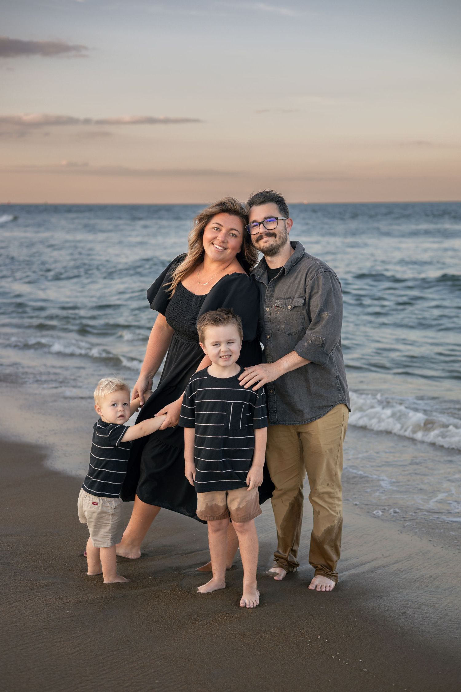 Family of four poses on beach at sunset, two young boys in striped shirts with parents in casual attire, ocean waves behind them.