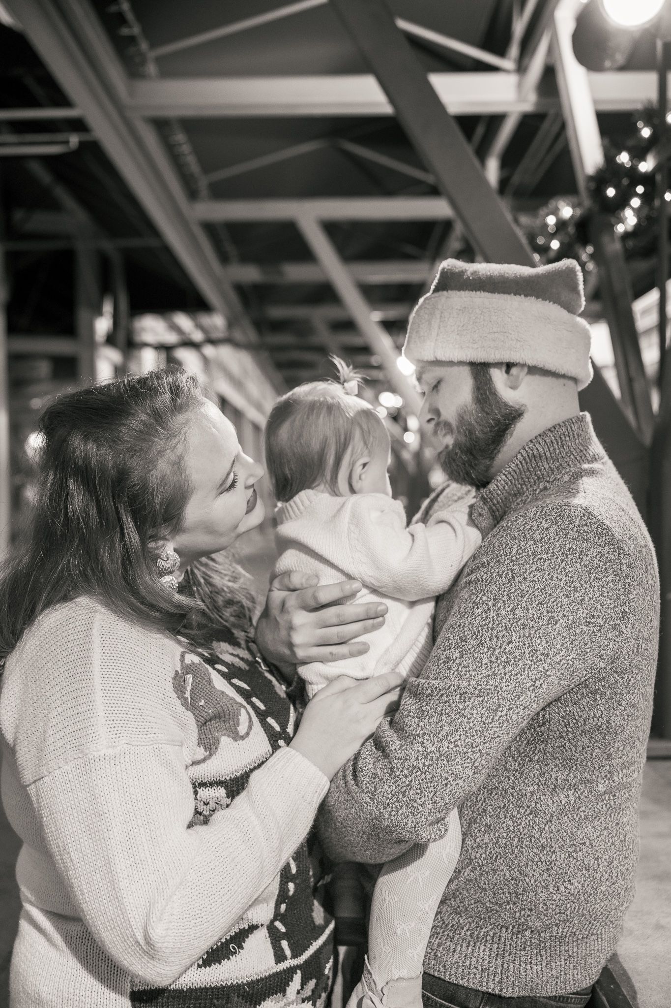 Black and white photo of a couple holding their baby, smiling warmly, with festive lights in the background.