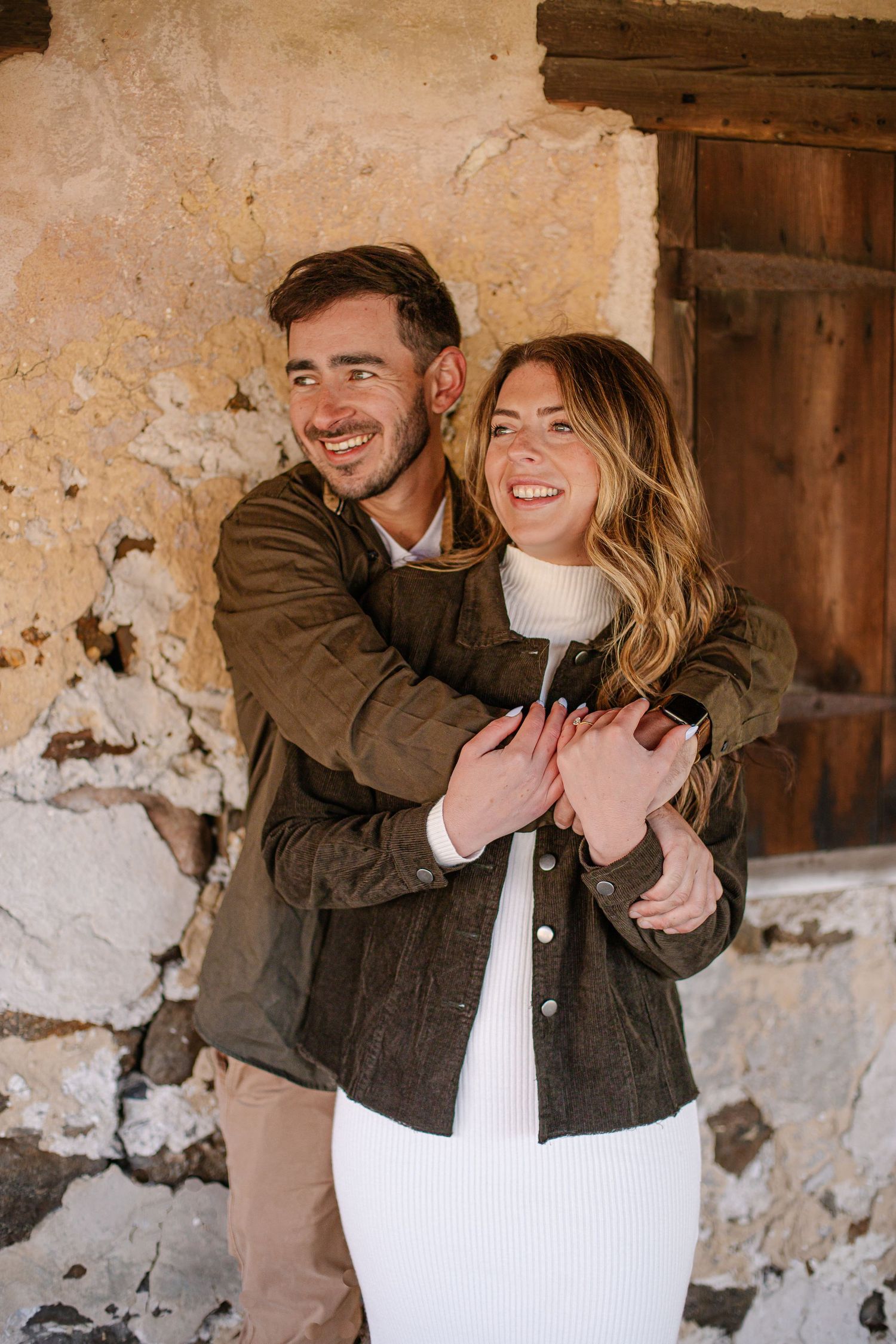 A smiling couple embraces against a rustic stone wall, both wearing olive green jackets, as the man hugs the woman from behind.
