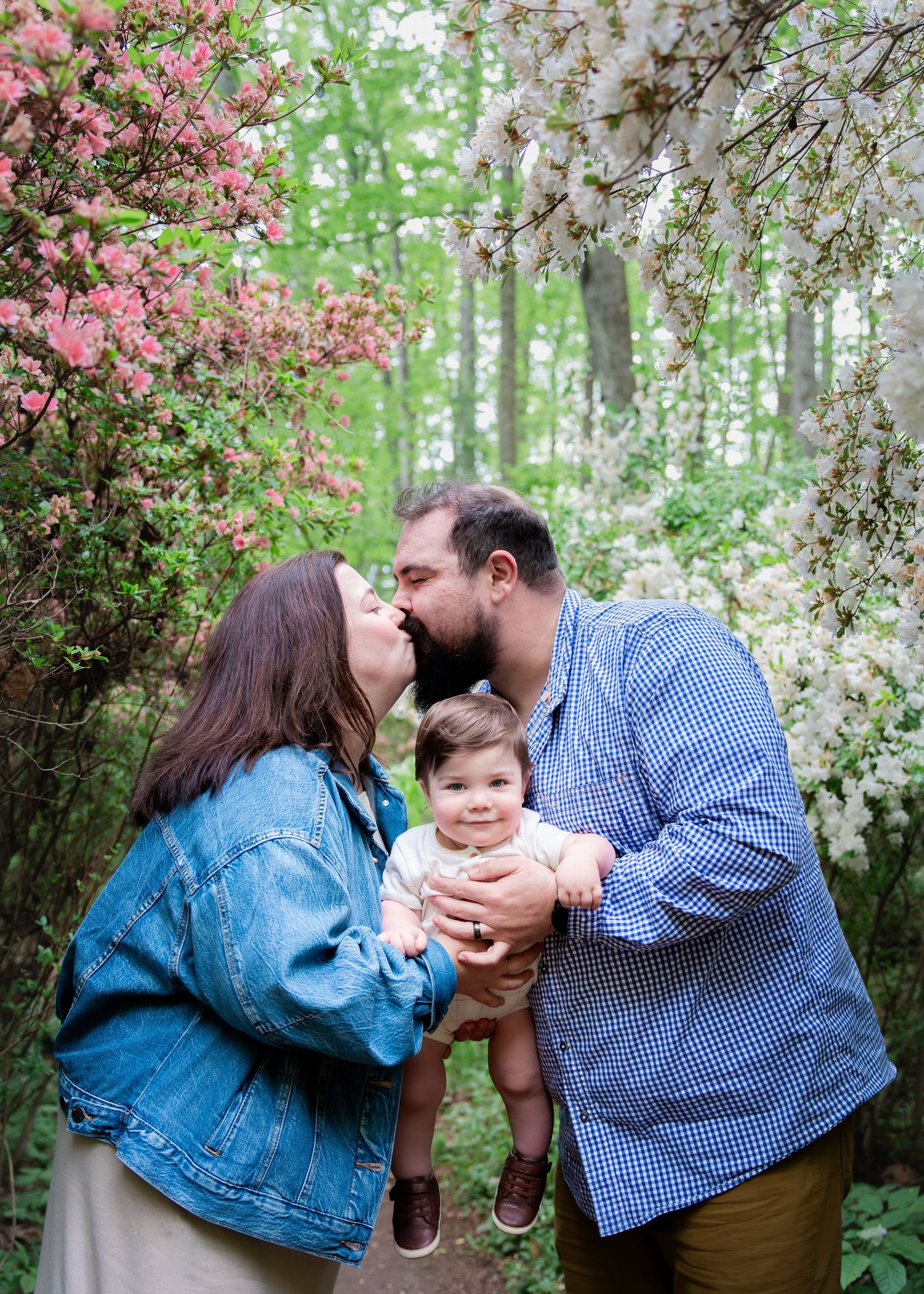 A couple kisses while holding their smiling baby amid blooming pink and white spring trees in a lush garden setting.