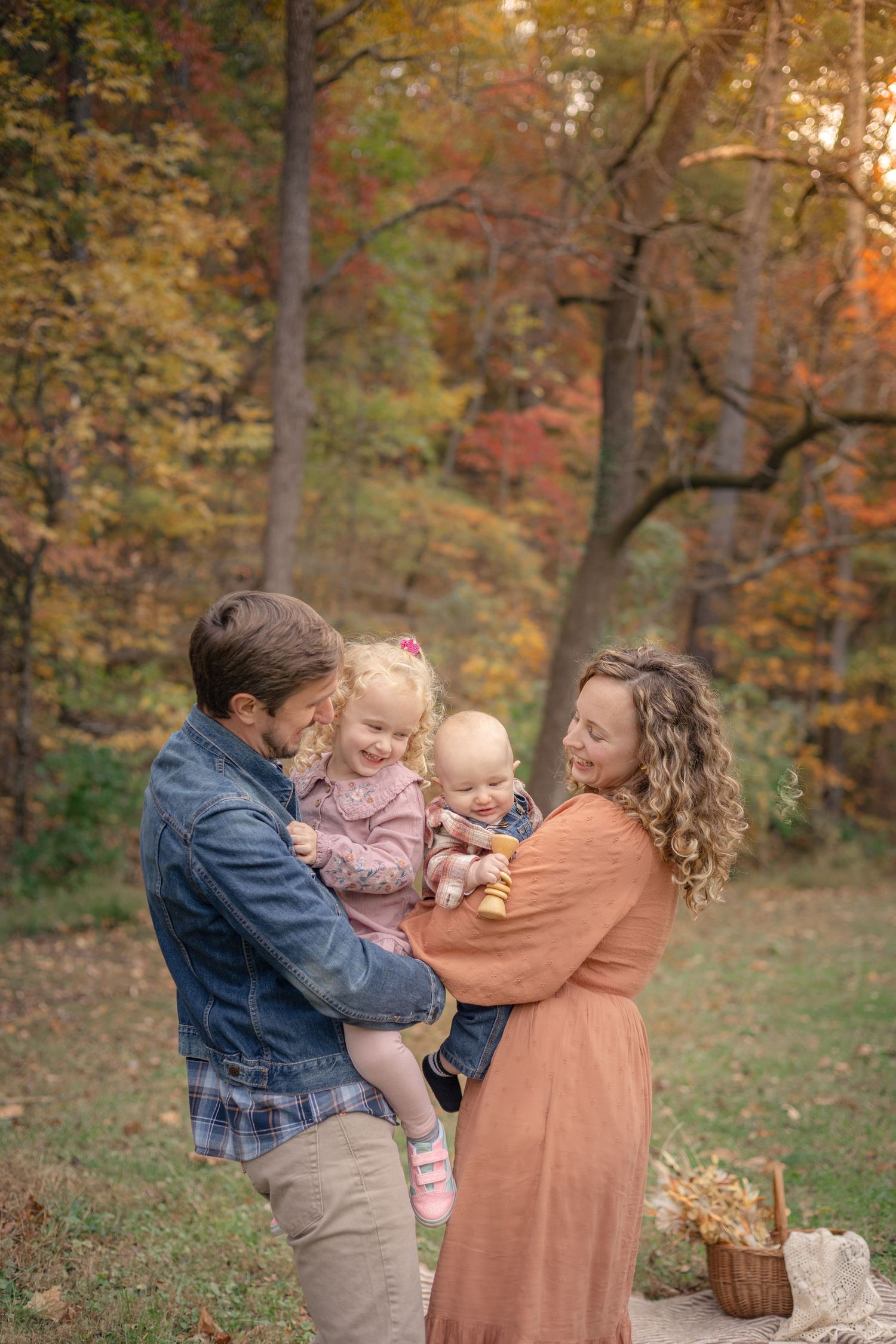 Family of four enjoying autumn foliage outdoors, parents holding two young children, smiling warmly in a colorful fall forest setting.