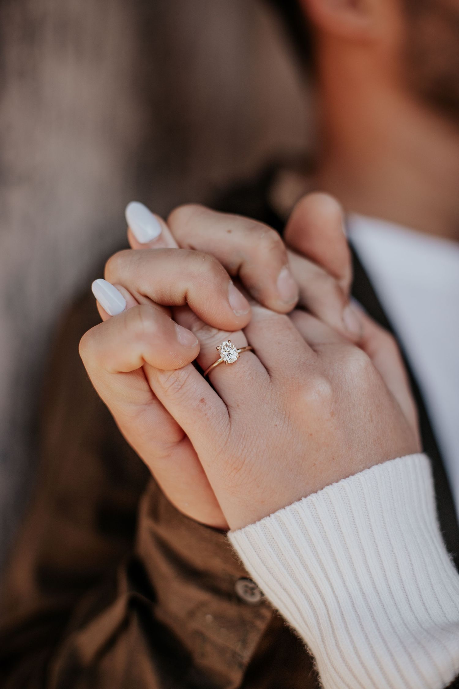 Close-up of intertwined hands showing a sparkling diamond engagement ring on a woman's finger with white manicured nails.