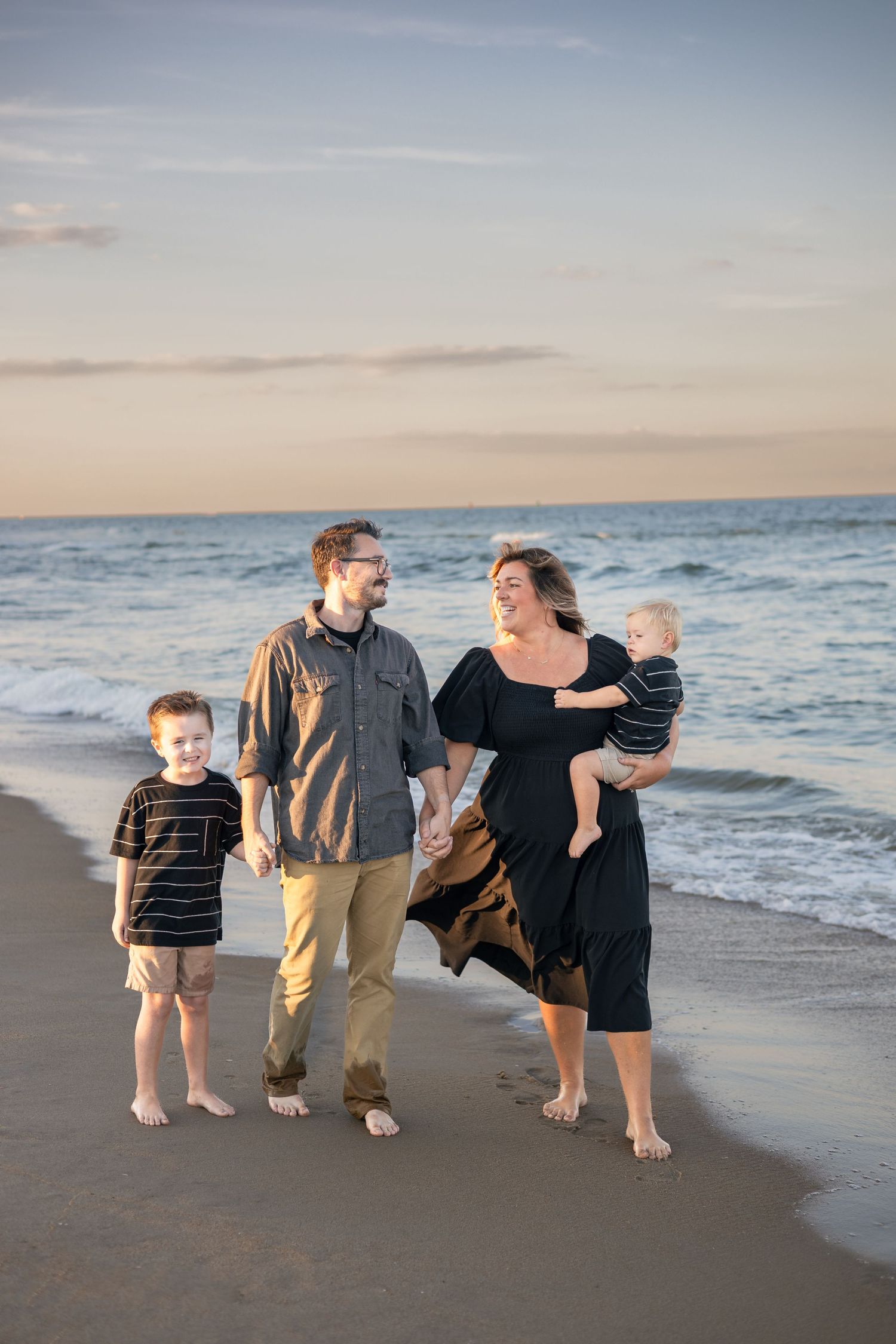 Family of four walking barefoot on a sandy beach at sunset, parents holding hands with two young boys.