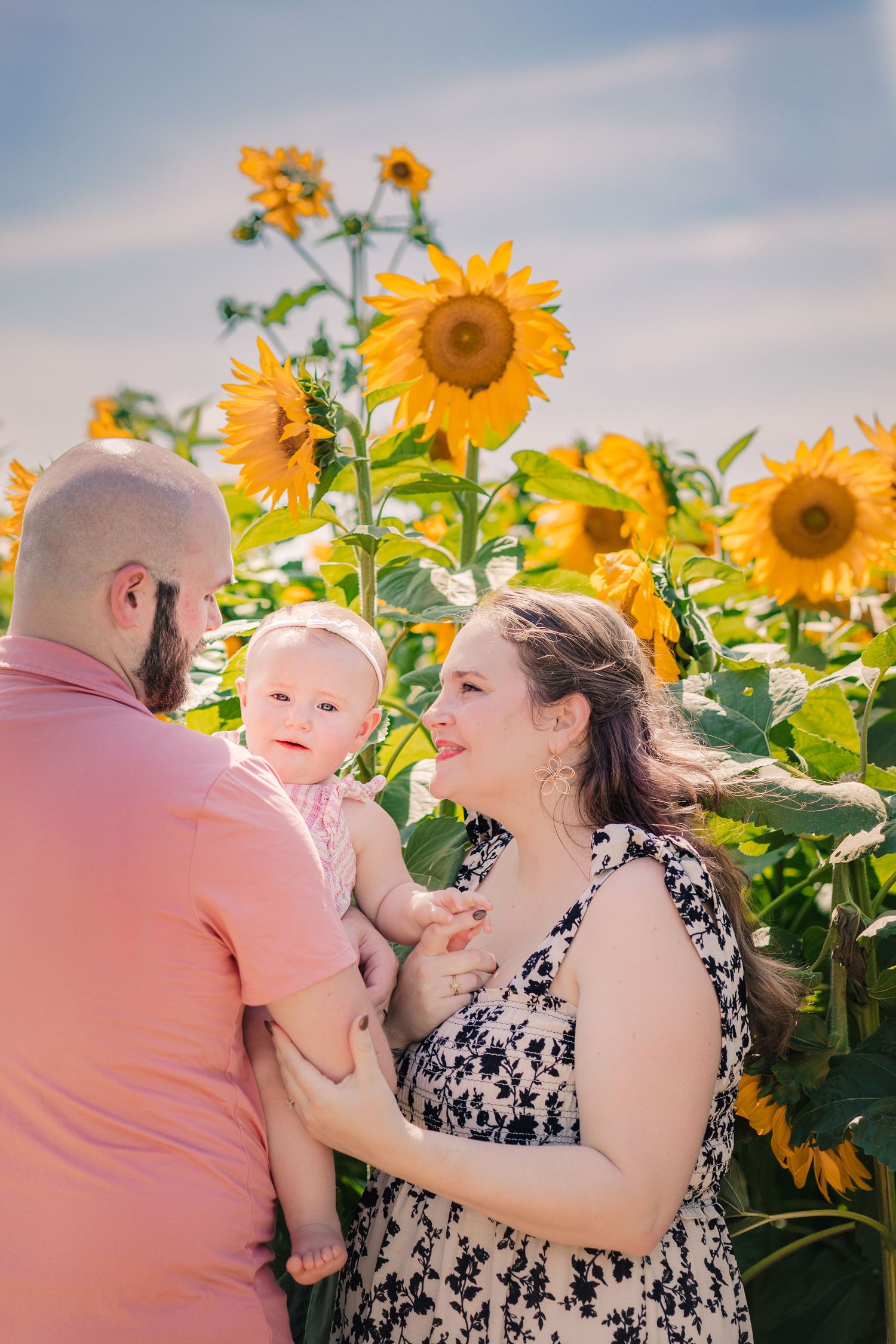 Family of three enjoying a sunny day in a vibrant sunflower field, parents lovingly gazing at their baby.
