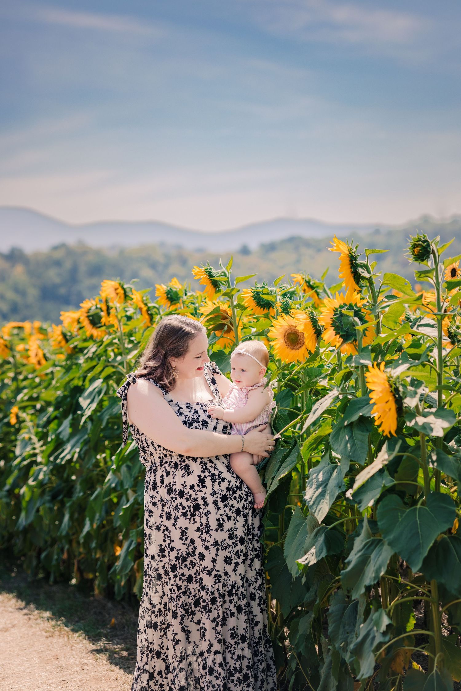 Mother in floral dress holds baby in sunflower field with mountains.