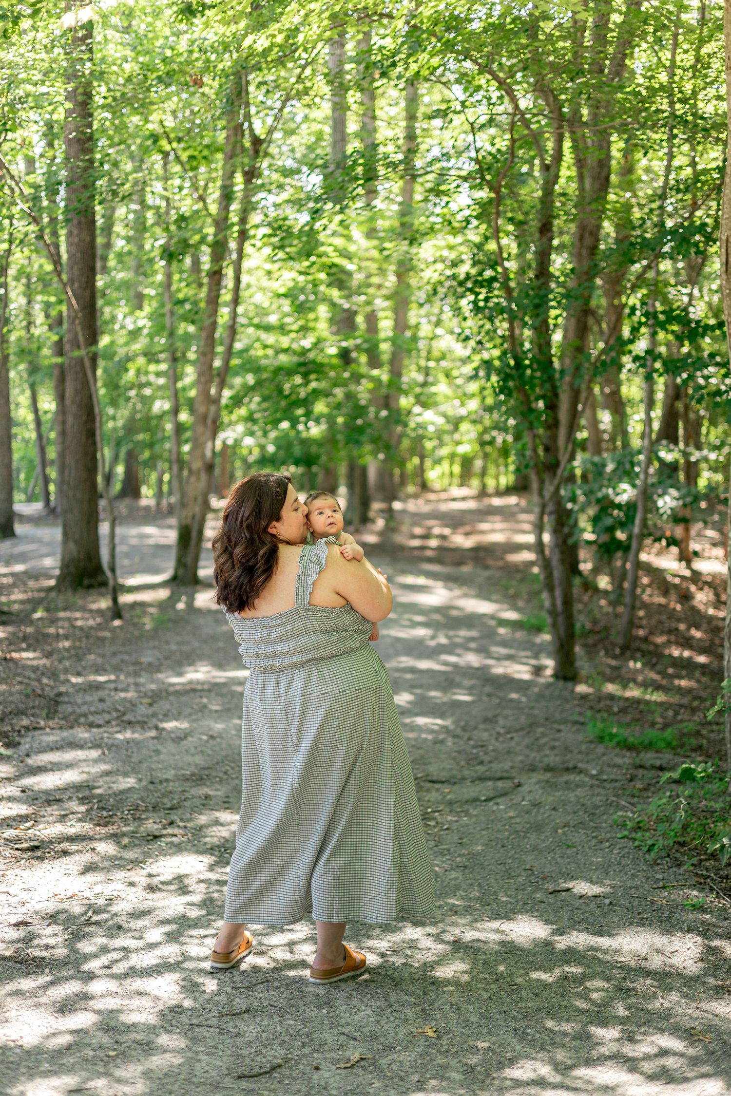 A mother in a sage green dress holds and kisses her baby on a sunlit forest path surrounded by tall trees.