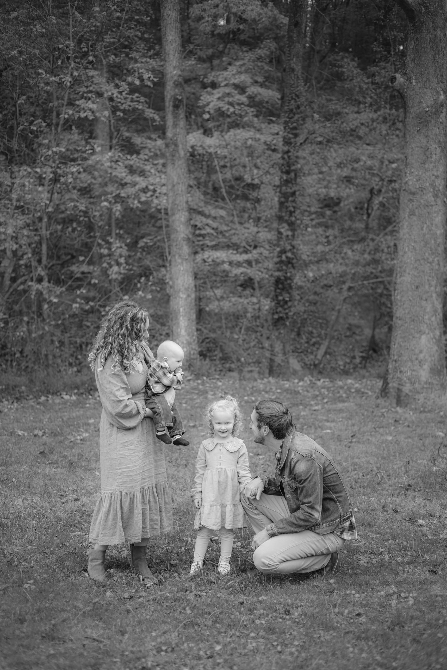Black and white family photo with parents and two young children enjoying time together in a wooded park setting.