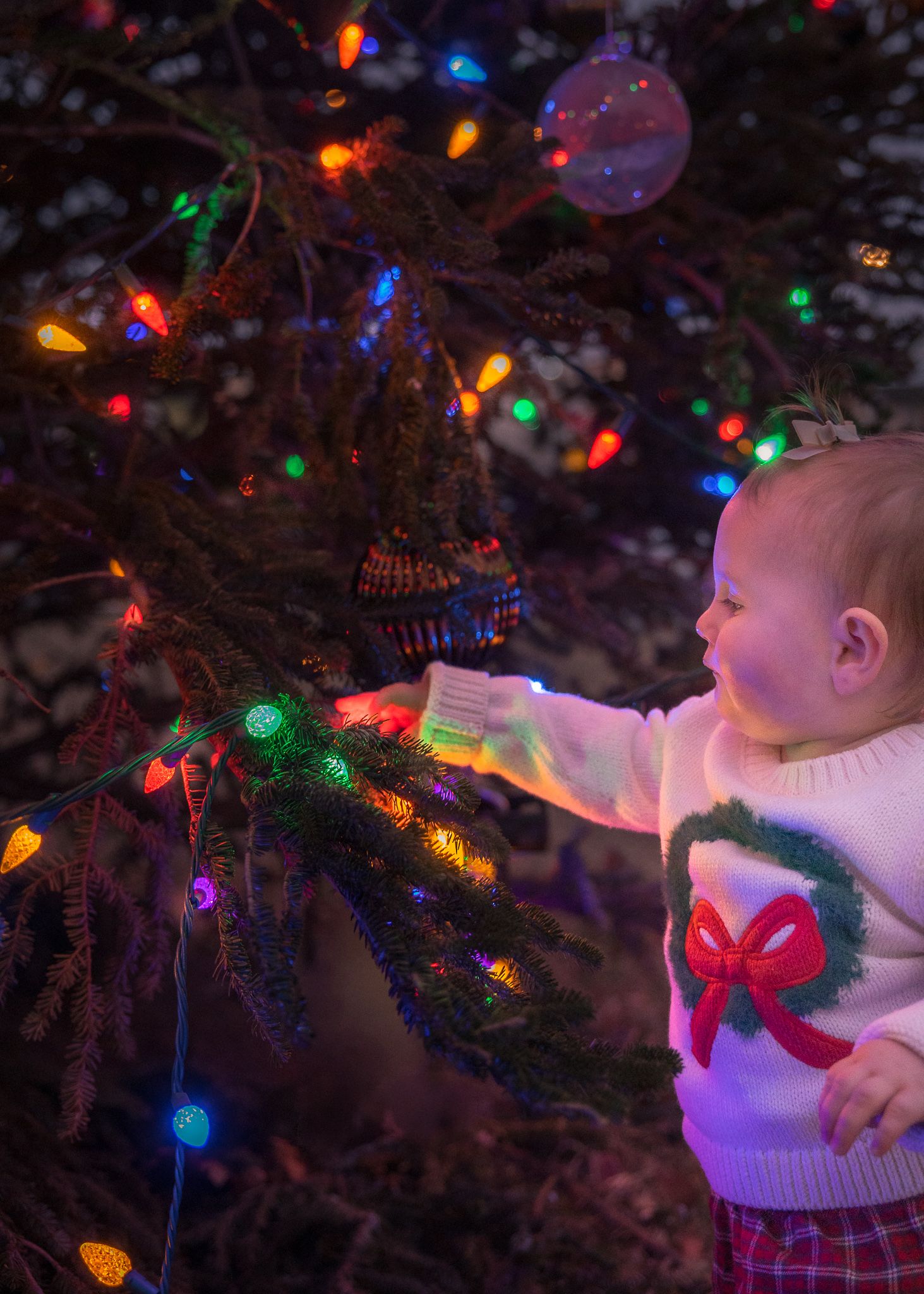 Baby in Christmas sweater reaching for colorful lights on decorated holiday tree.