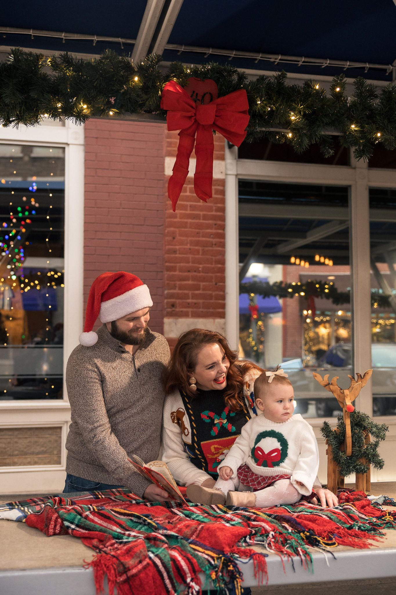 Family in Christmas sweaters and Santa hat with toddler, sitting under holiday wreath and lights at festive outdoor venue. Roanoke, VA