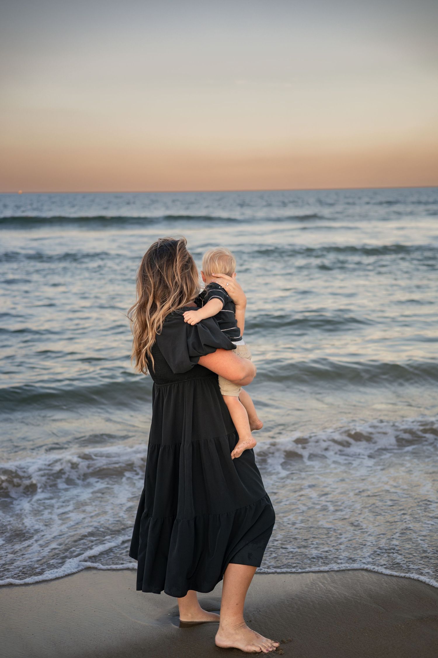 Woman in black dress holds baby at beach during golden hour sunset.