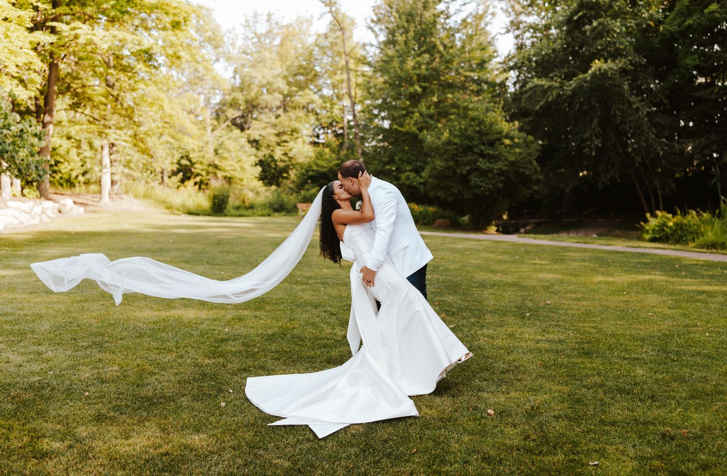 Flowing white wedding dress and veil catch the breeze in sunny garden setting.