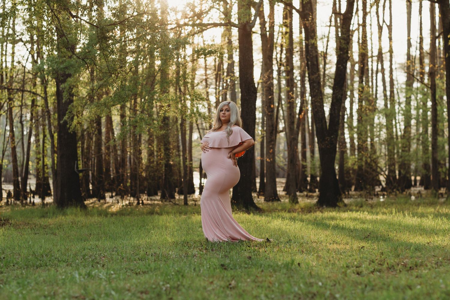 A person in a flowing pink dress poses in a sunlit forest setting during golden hour.