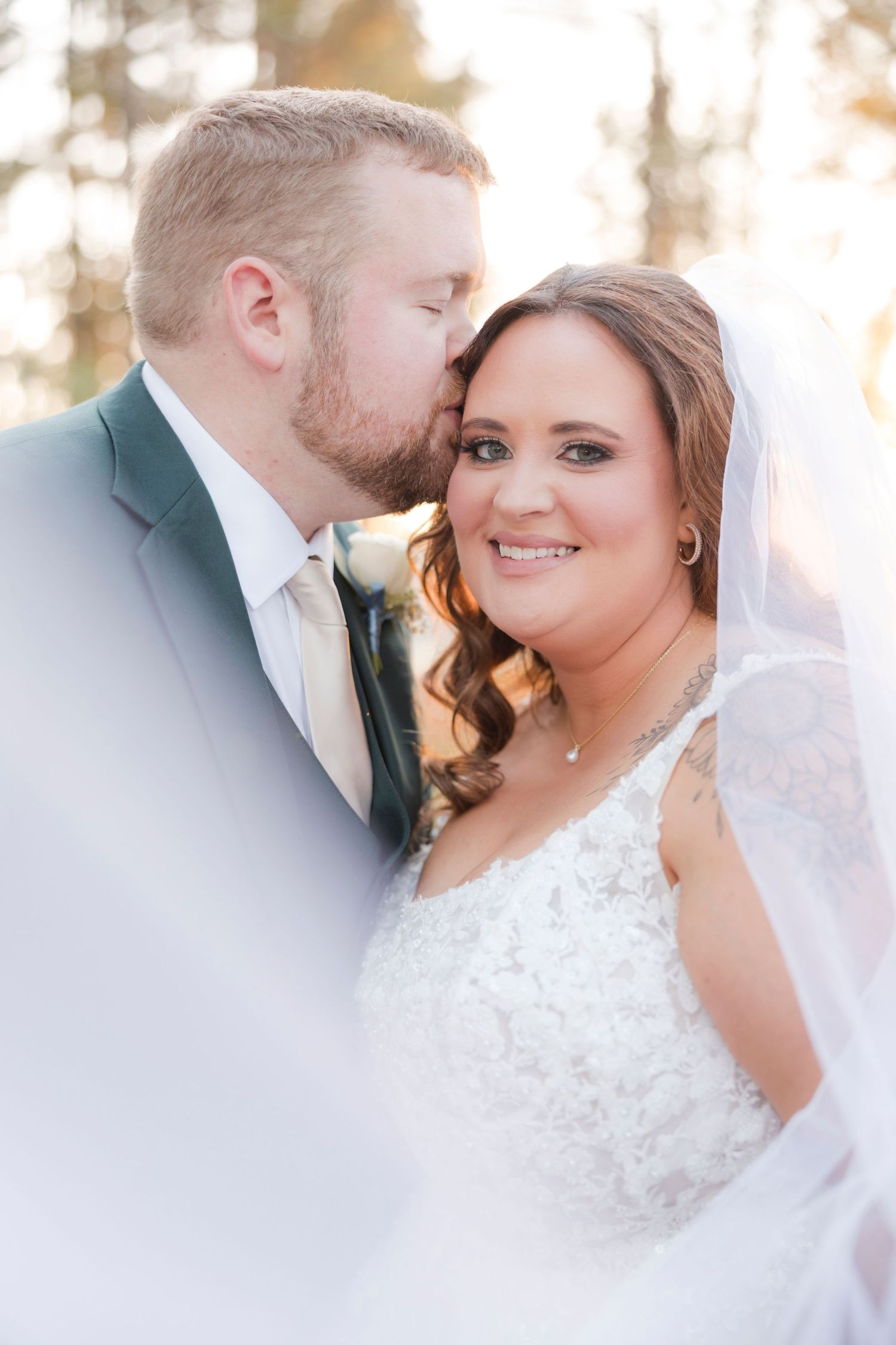 Groom in gray suit kisses bride on cheek as she smiles in white lace gown and veil on wedding day.