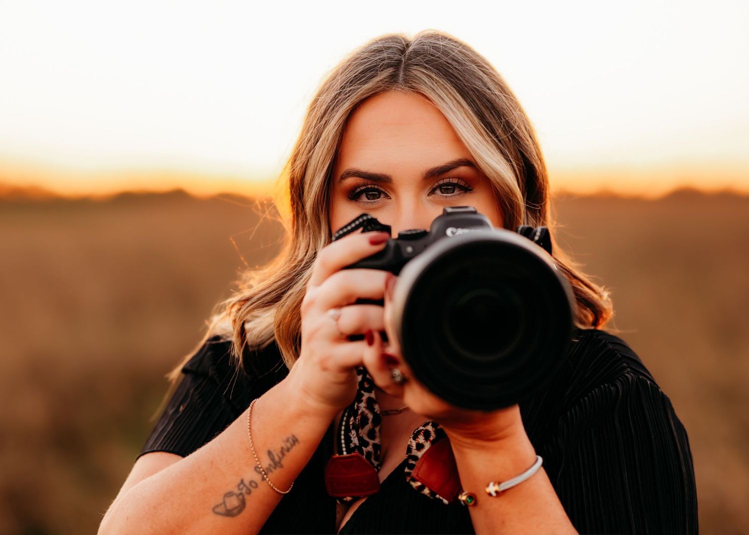 A photographer with long blonde hair holds a professional camera against a warm sunset background in an open field.