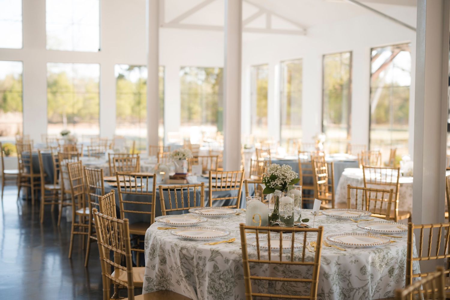 Elegant wedding reception venue with gold Chiavari chairs, floral tablecloths, and large windows overlooking a garden.