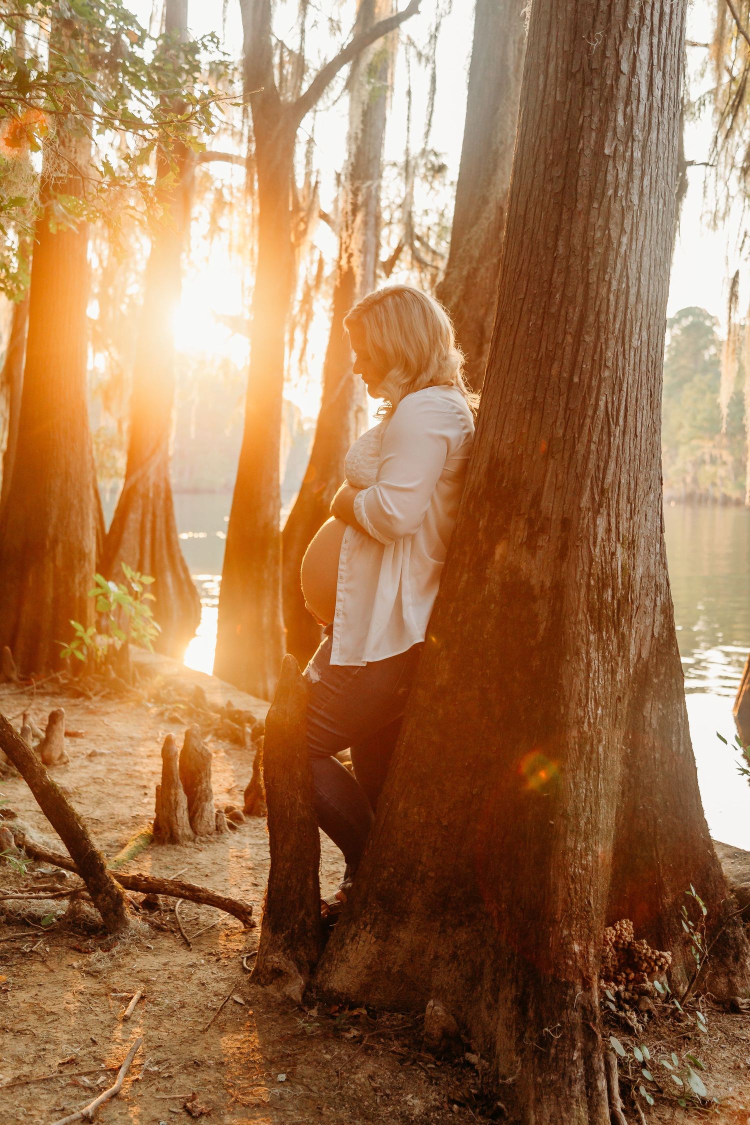 A silhouette leans against a cypress tree at sunset by a lake with golden sunlight filtering through the trees.