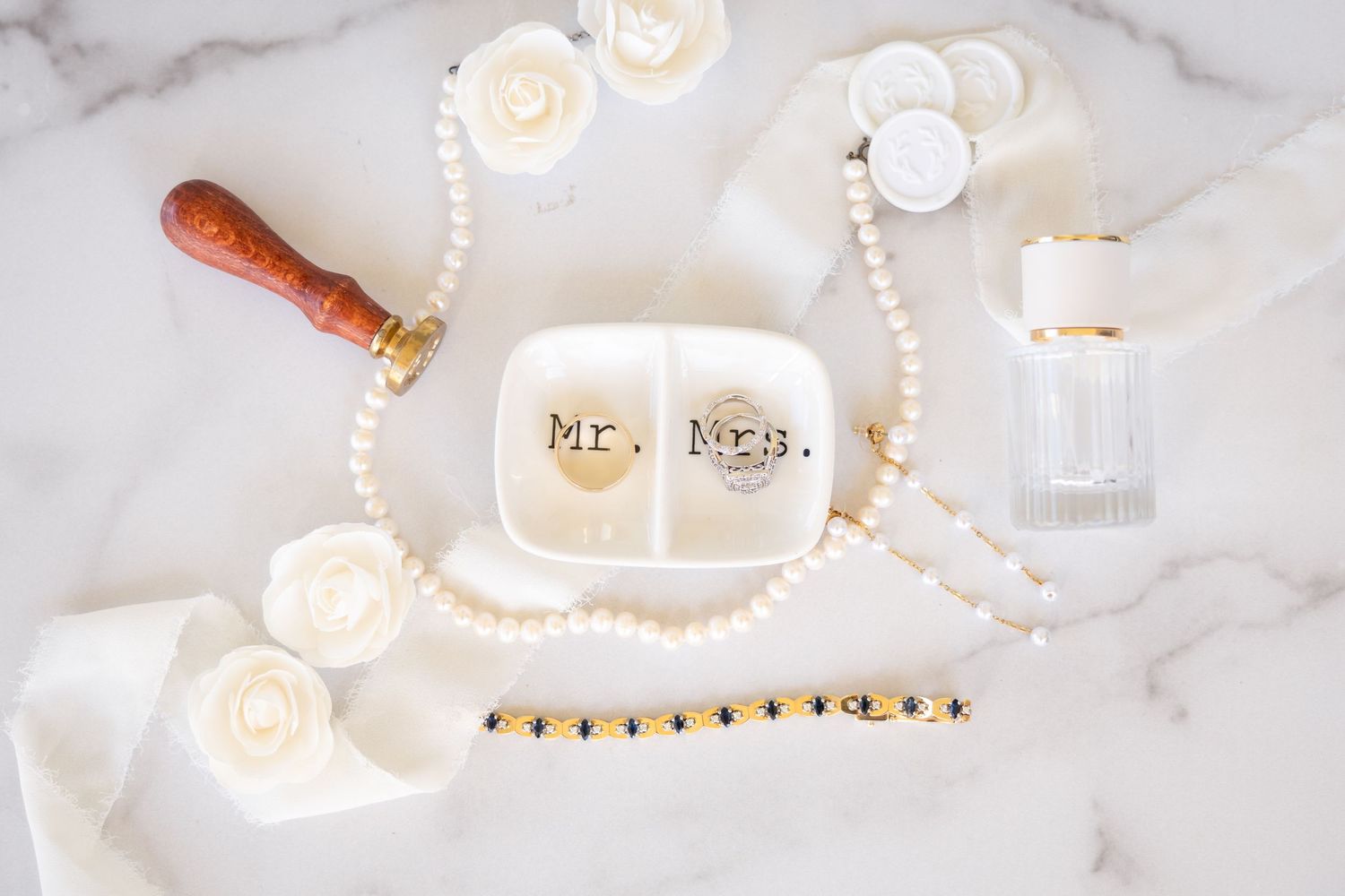 Mr. and Mrs. ceramic dish with pearl necklace, wax seal, roses, and crystal perfume bottle on marble surface.
