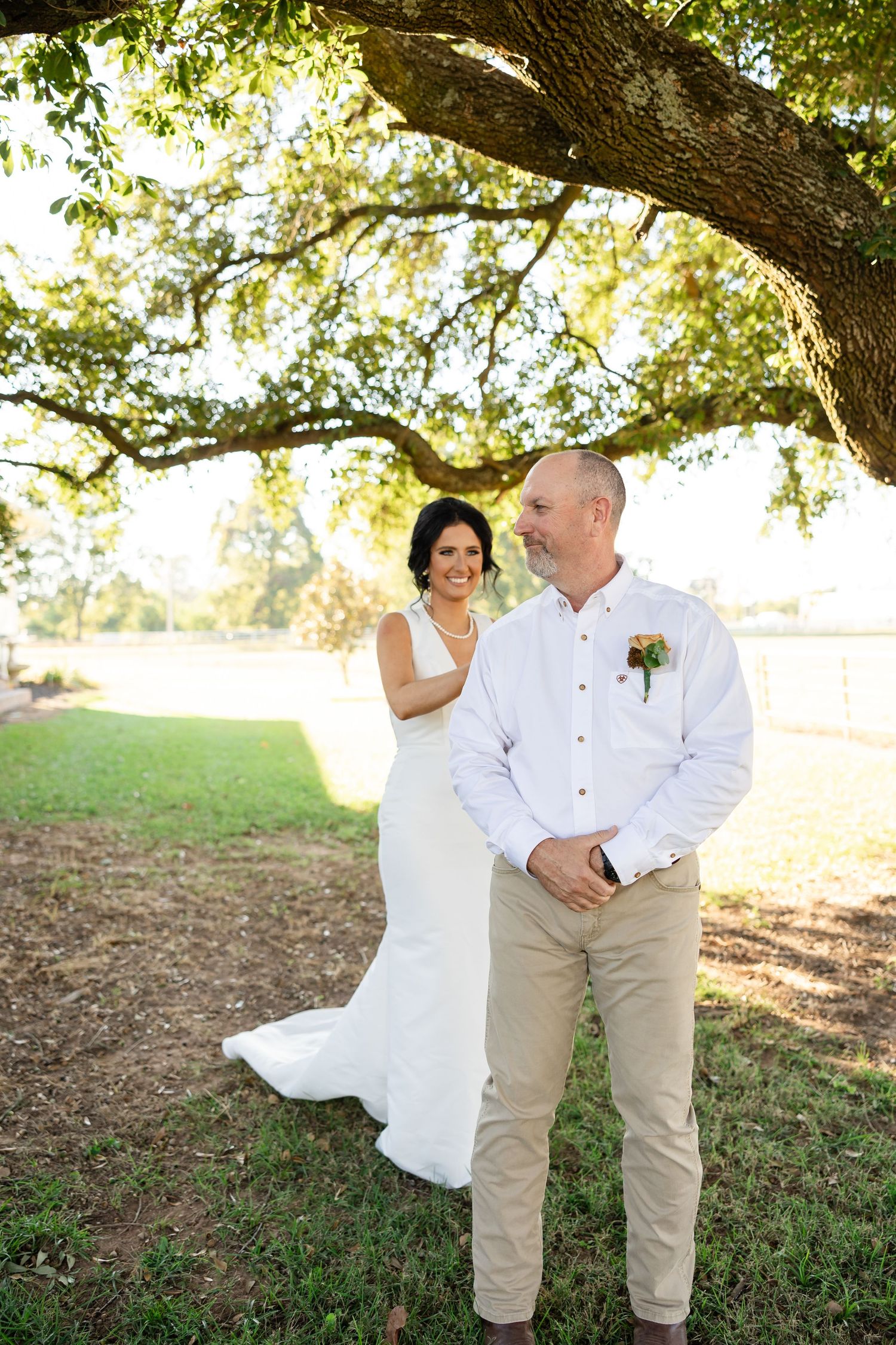 Bride in white gown and groom in white shirt with boutonniere pose under large oak tree at outdoor wedding.