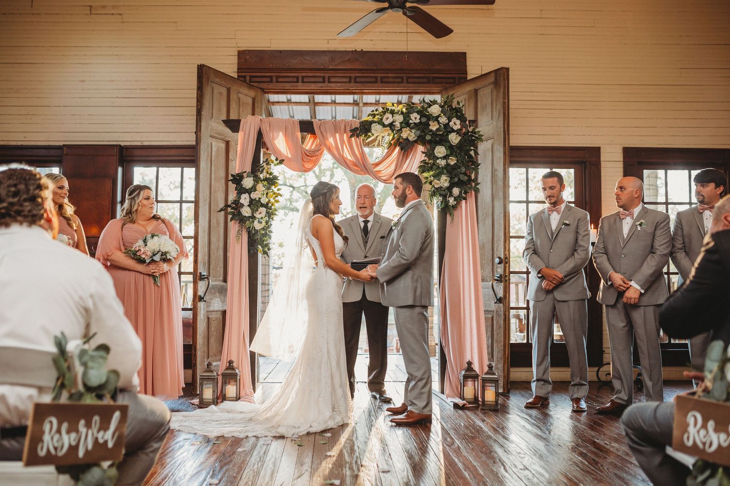 Couple exchanging vows at rustic indoor wedding ceremony with floral arch, pink drapes, and groomsmen in gray suits.