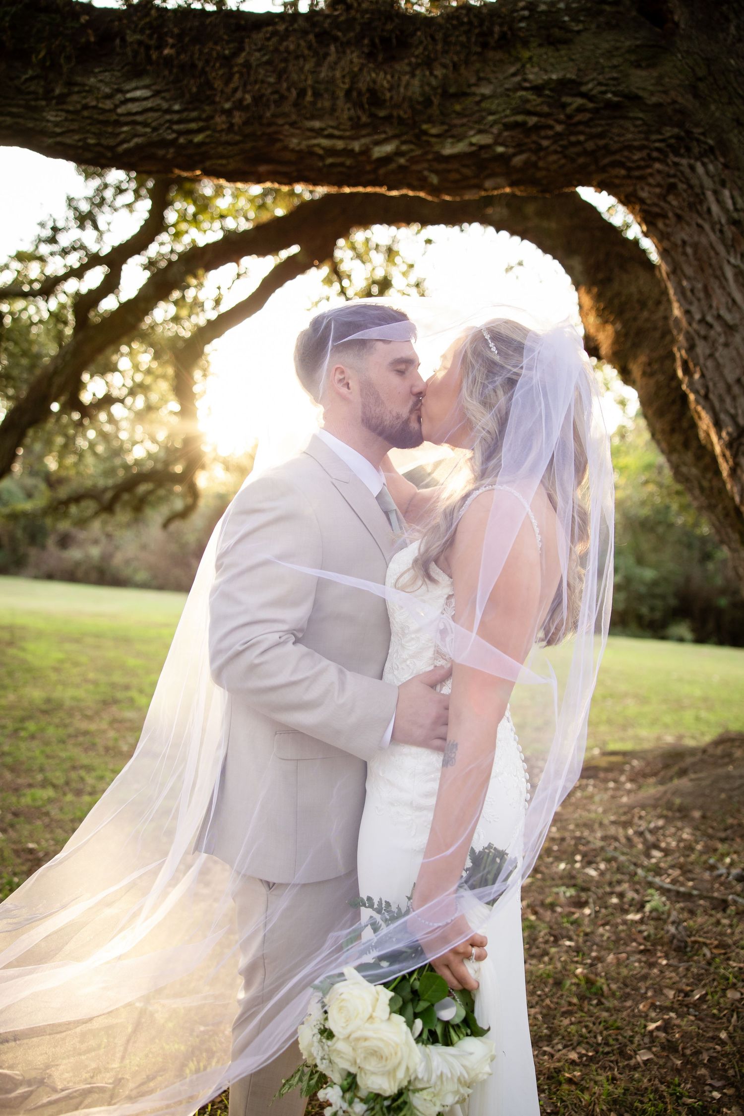 Bride and groom share a romantic kiss under a sprawling oak tree at golden hour, veil flowing in the breeze.