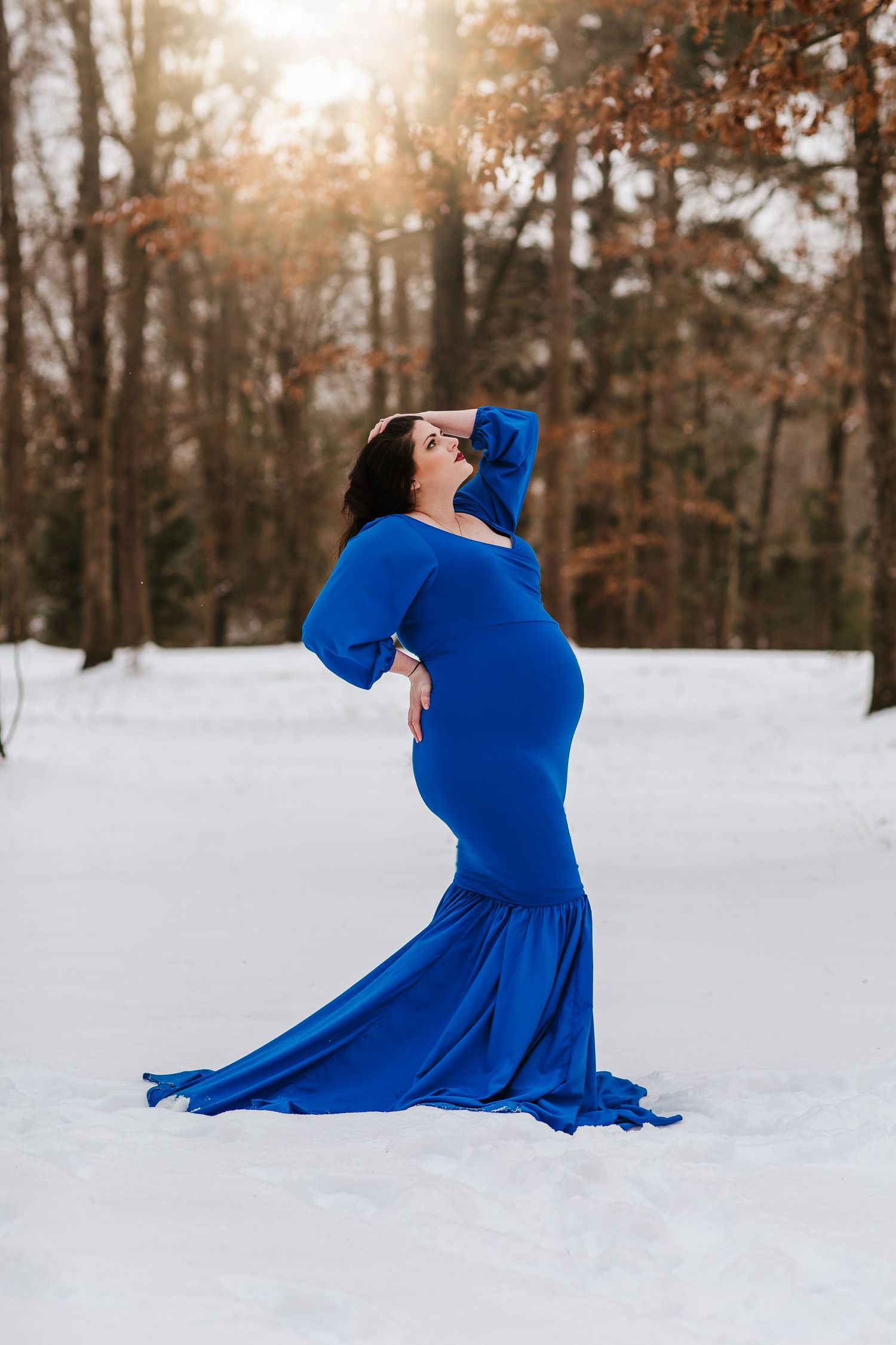 A woman in a flowing royal blue dress poses gracefully in a snowy forest during winter as sunlight filters through trees.