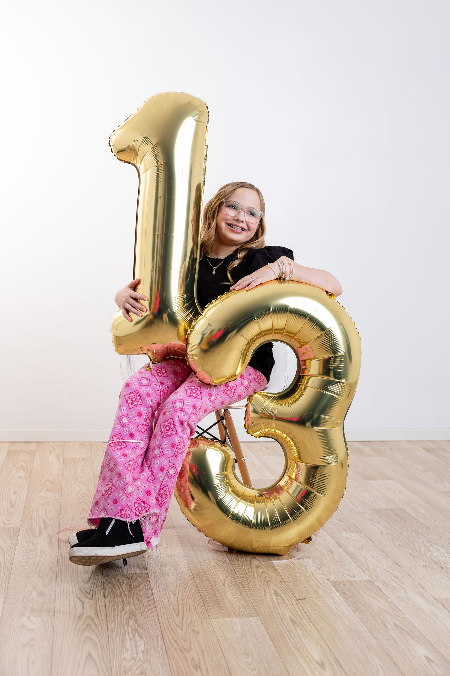 A person sits with large gold balloon numbers 13 against a white wall on hardwood flooring.