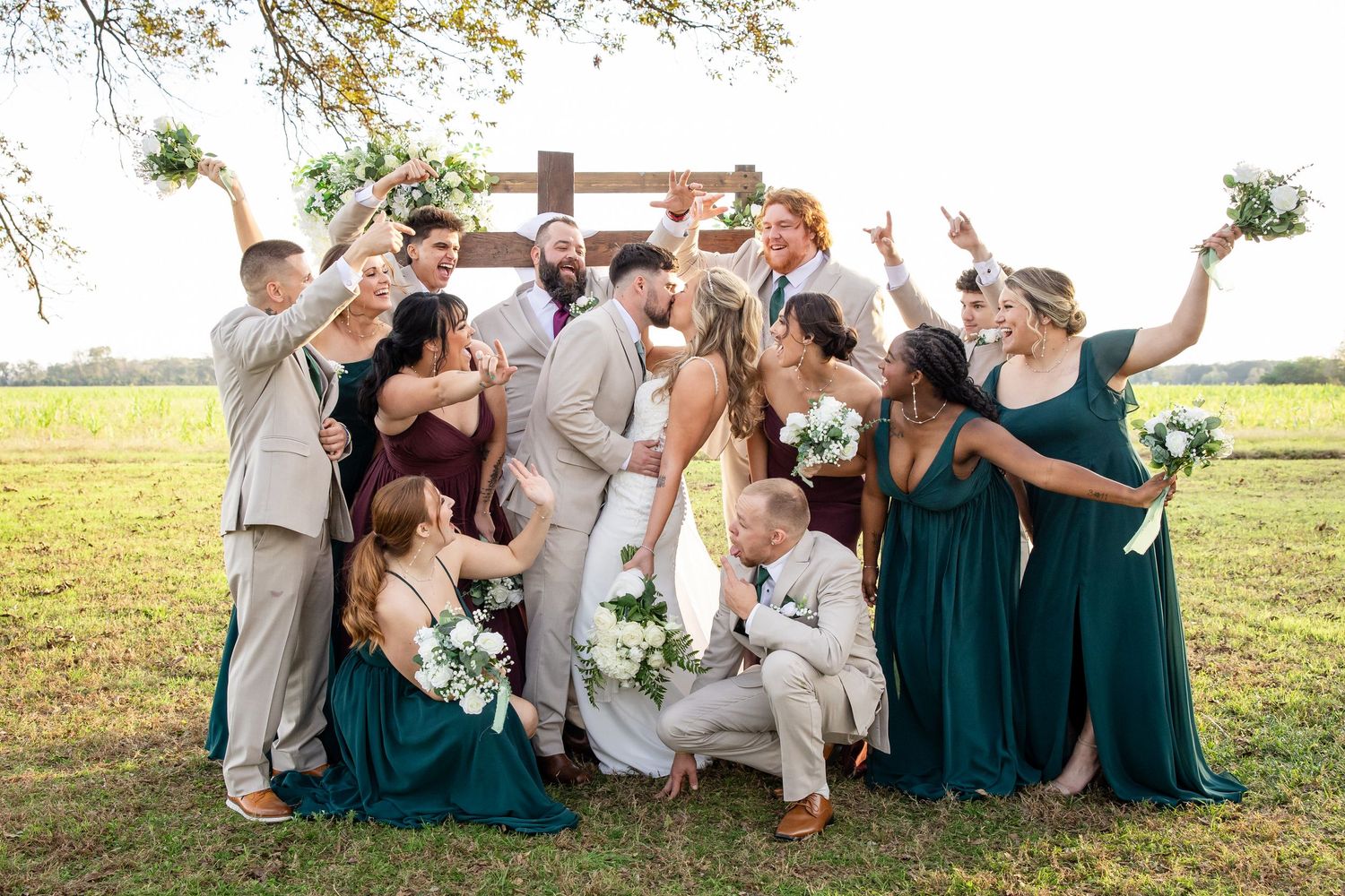 Wedding party in teal dresses and khaki suits sits together on grassy lawn during outdoor celebration.