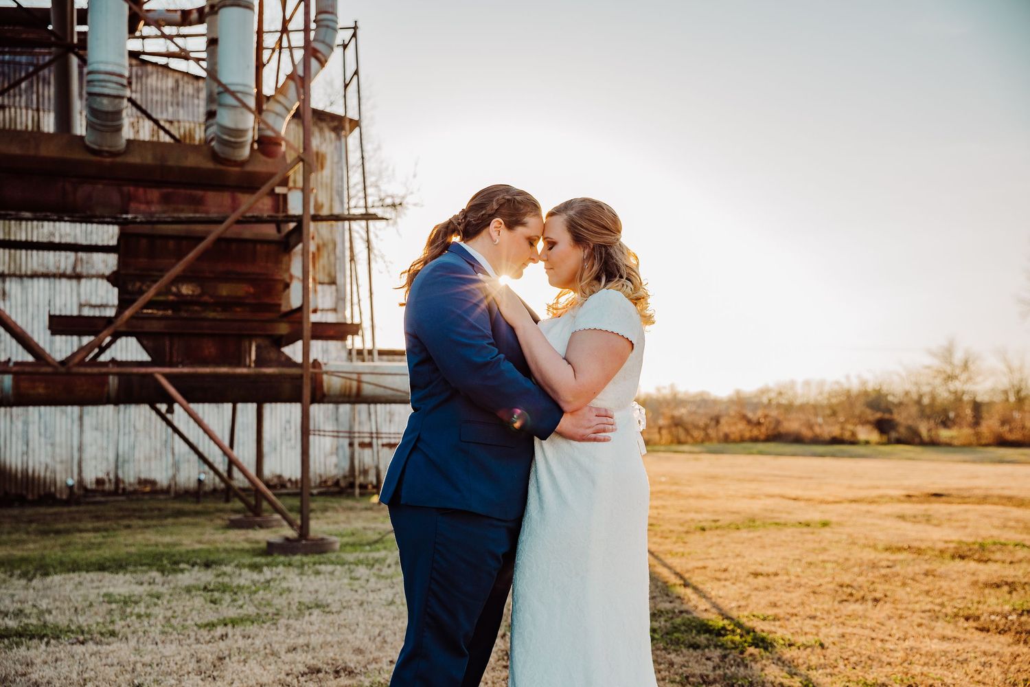 A couple shares an intimate kiss in a rustic outdoor setting with golden sunlight and farm buildings in the background.