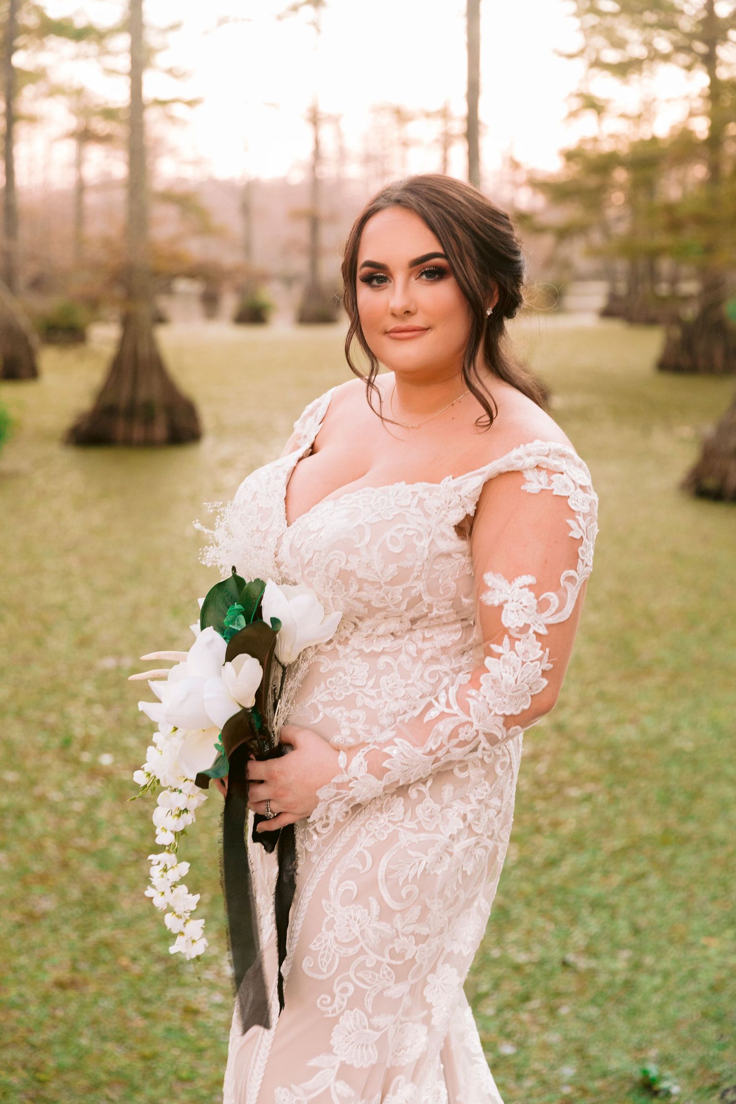 Beautiful bridal portrait in a lace white wedding dress holding white flowers in a misty cypress grove at sunset.