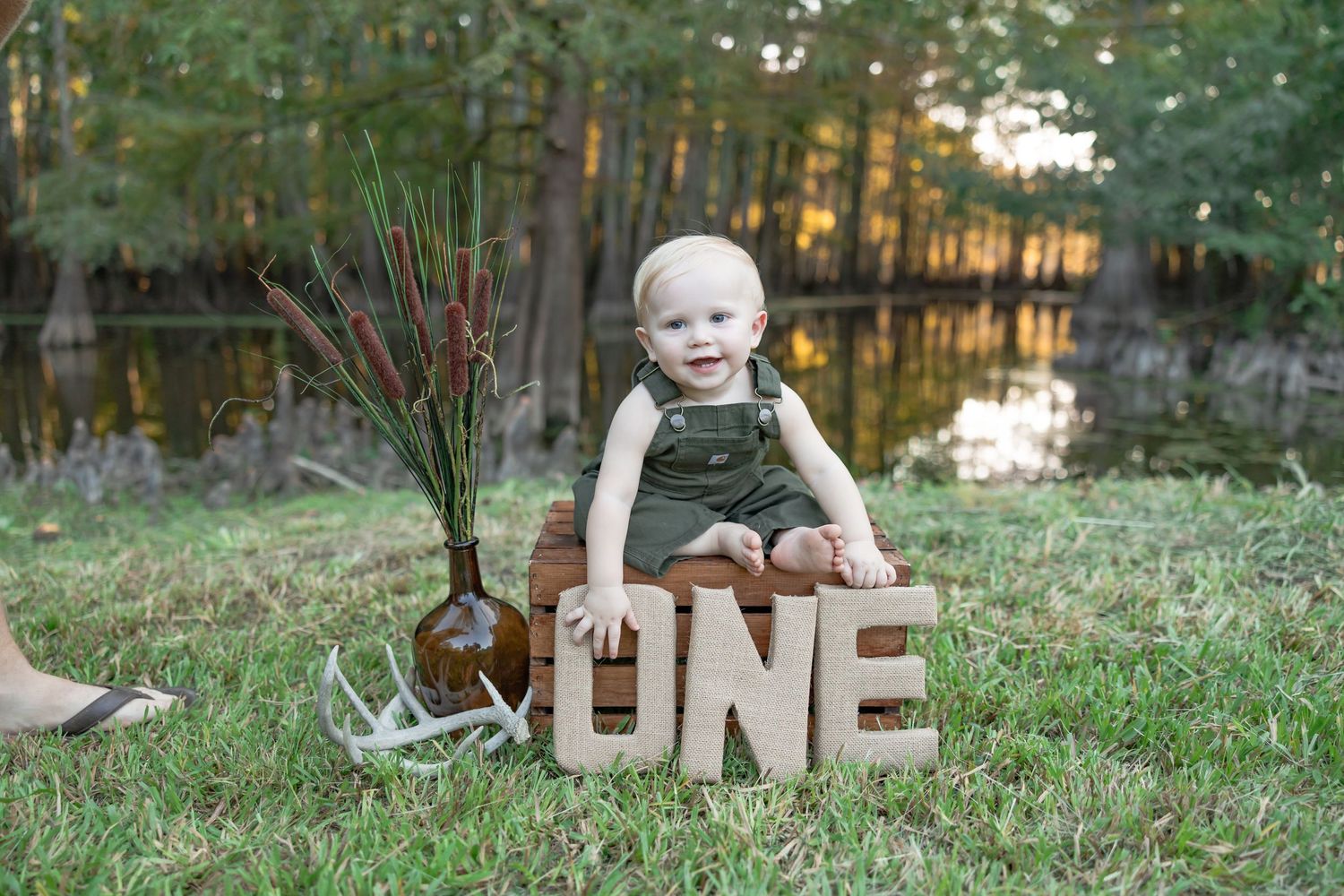 A small child sits on grass by a lake with wooden ONE letters and a rustic antler decoration for a first birthday photo.