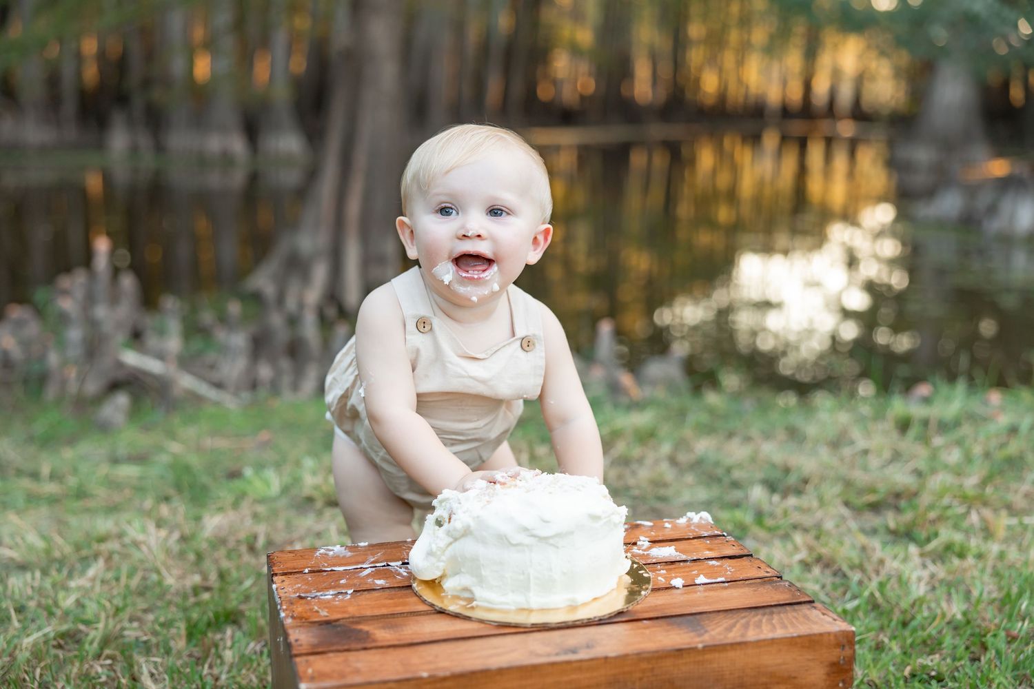 A smiling child in tan overalls sits at a wooden table with a white birthday cake near a reflective pond.