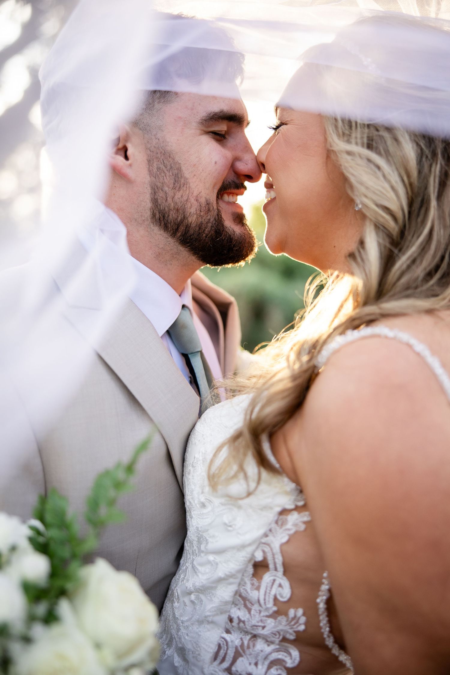 Bride and groom share a romantic almost-kiss under a flowing veil, dressed in lace gown and tan suit.
