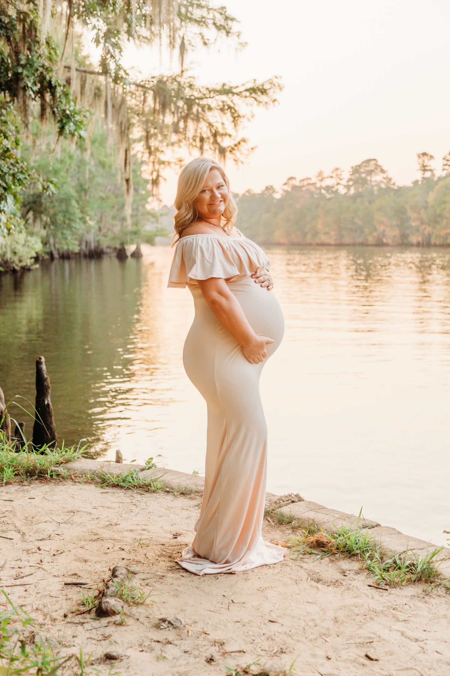 A pregnant woman in a white off-shoulder dress poses by a tranquil lake at sunset with Spanish moss trees in the background.