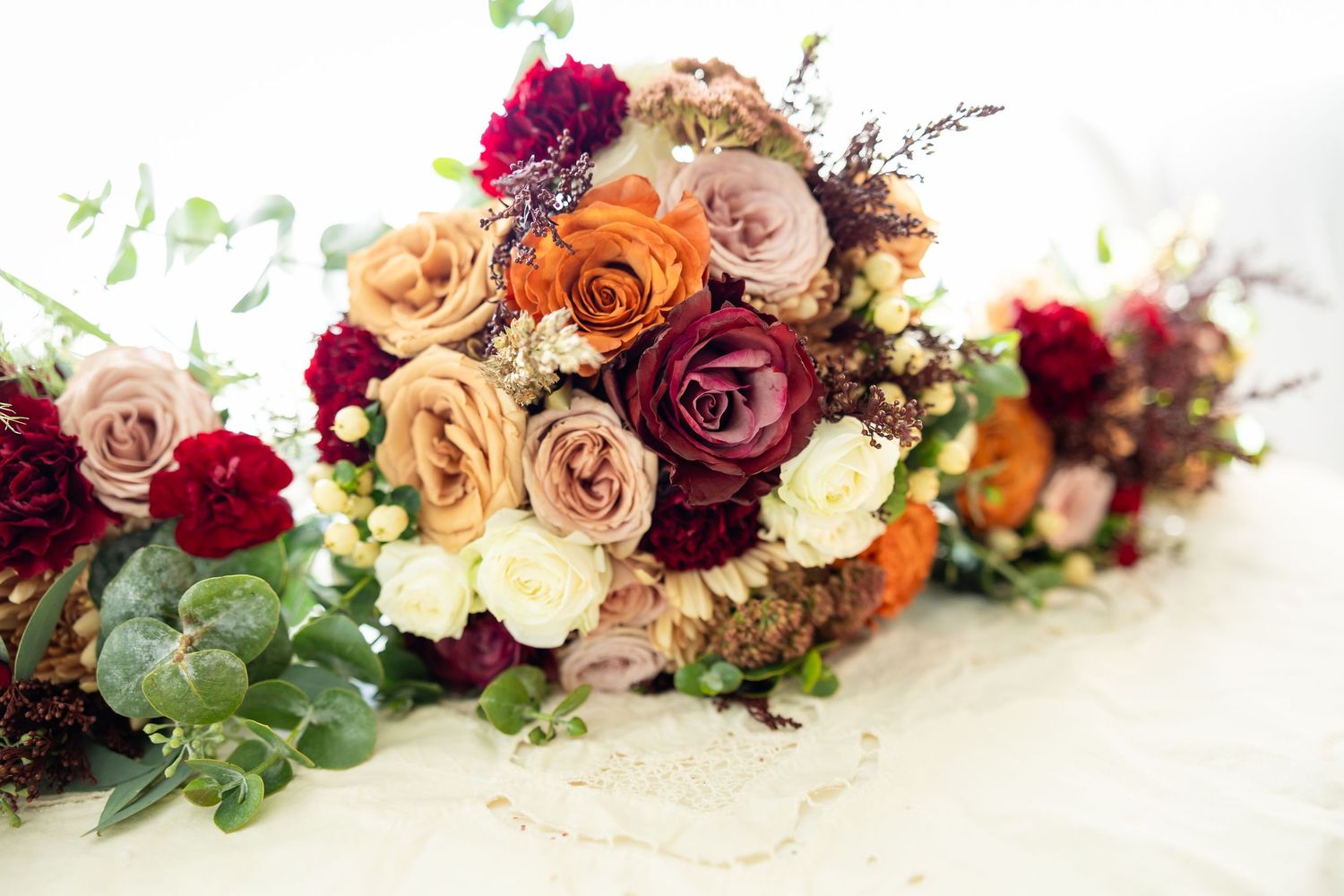 Lush wedding bouquets featuring burgundy, blush, and orange roses with eucalyptus greenery arranged on a white surface.