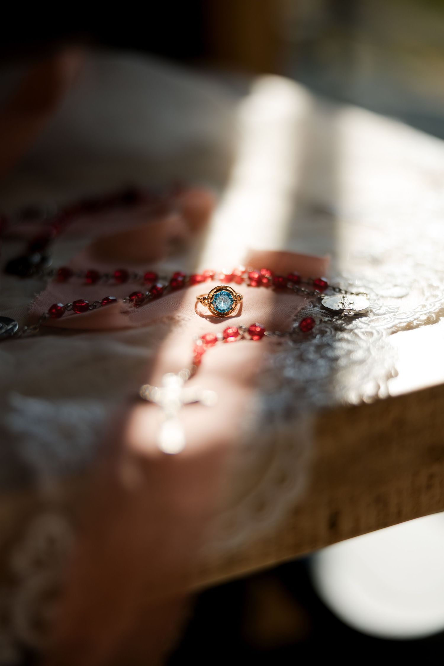 Gold ring with turquoise gemstone beside red rosary beads on wooden surface in warm sunlight.