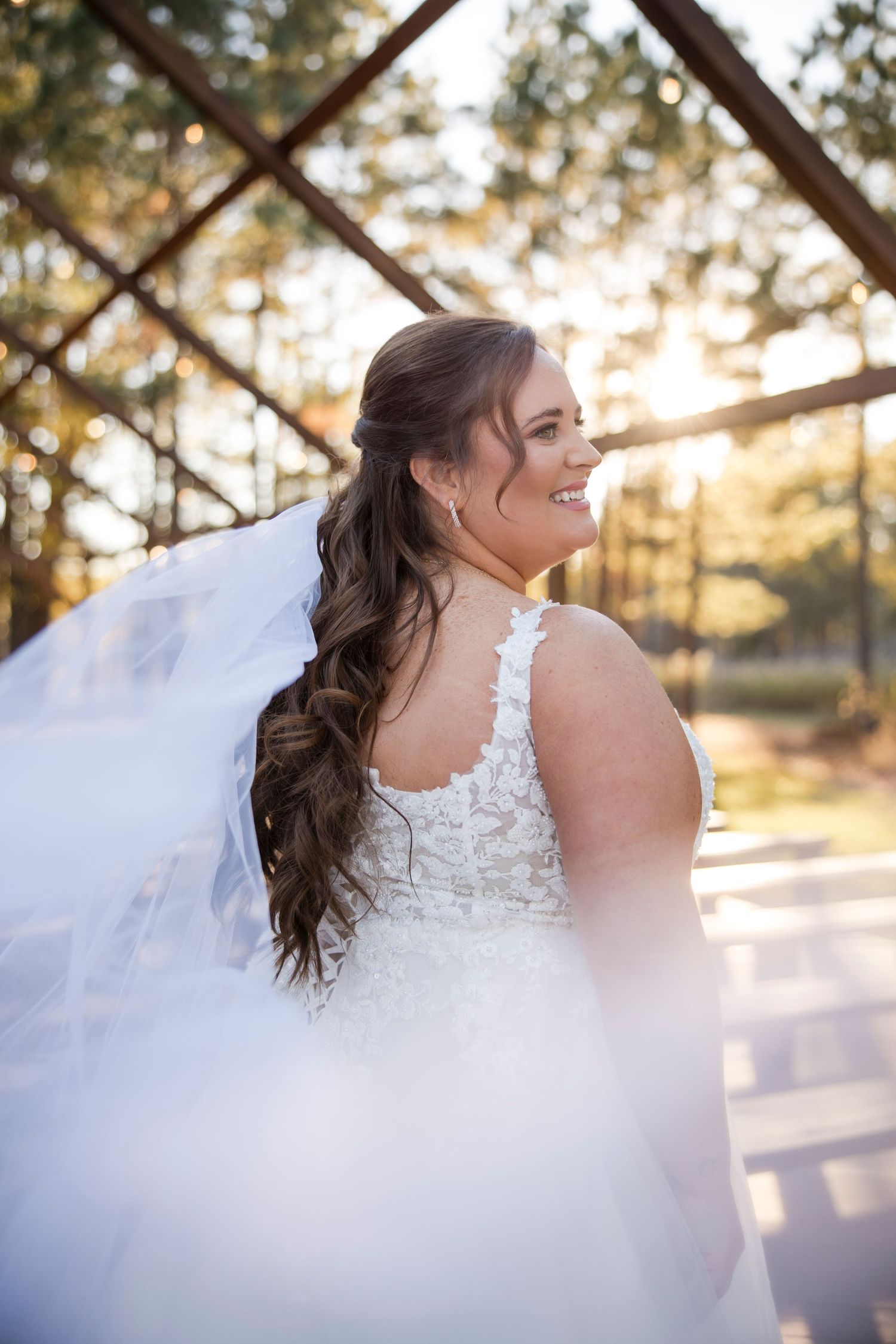 Smiling bride in lace wedding gown with flowing veil, long wavy hair, posing outdoors in golden sunset light.