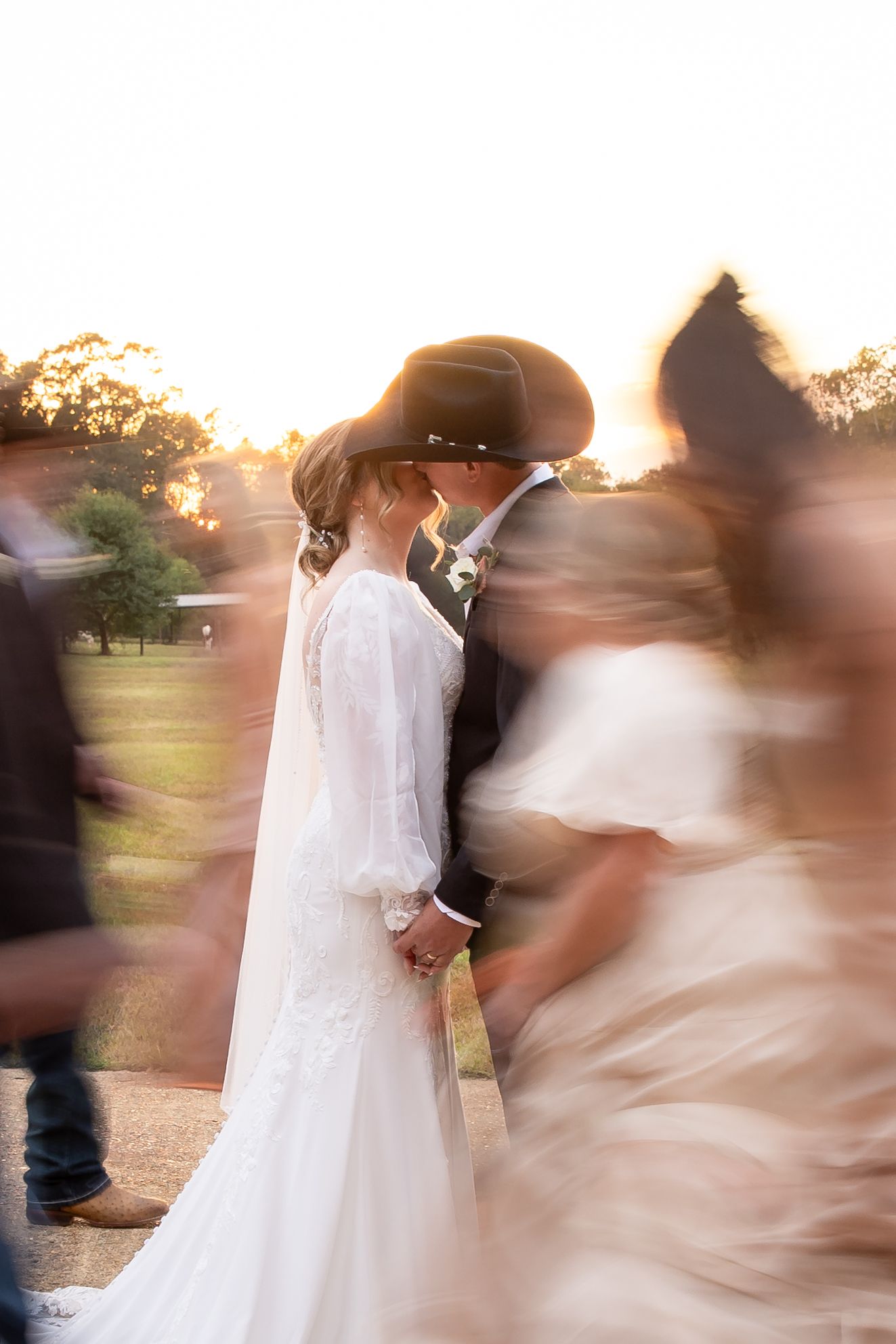 Bride and groom kissing at sunset in cowboy hats, white lace dress, with blurred wedding guests moving around them.