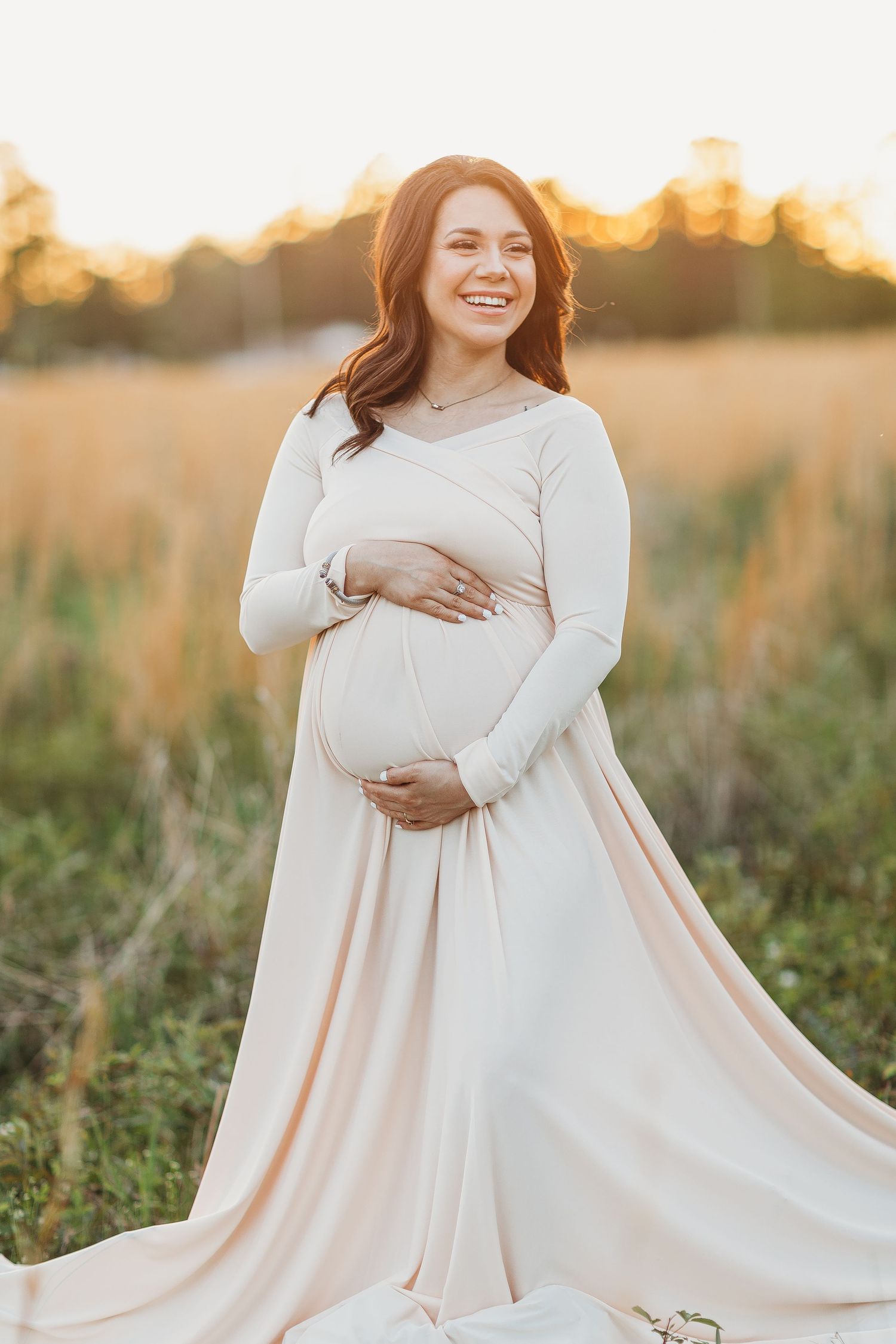 Expectant woman in flowing white dress poses in golden sunlit field during maternity photoshoot at sunset.