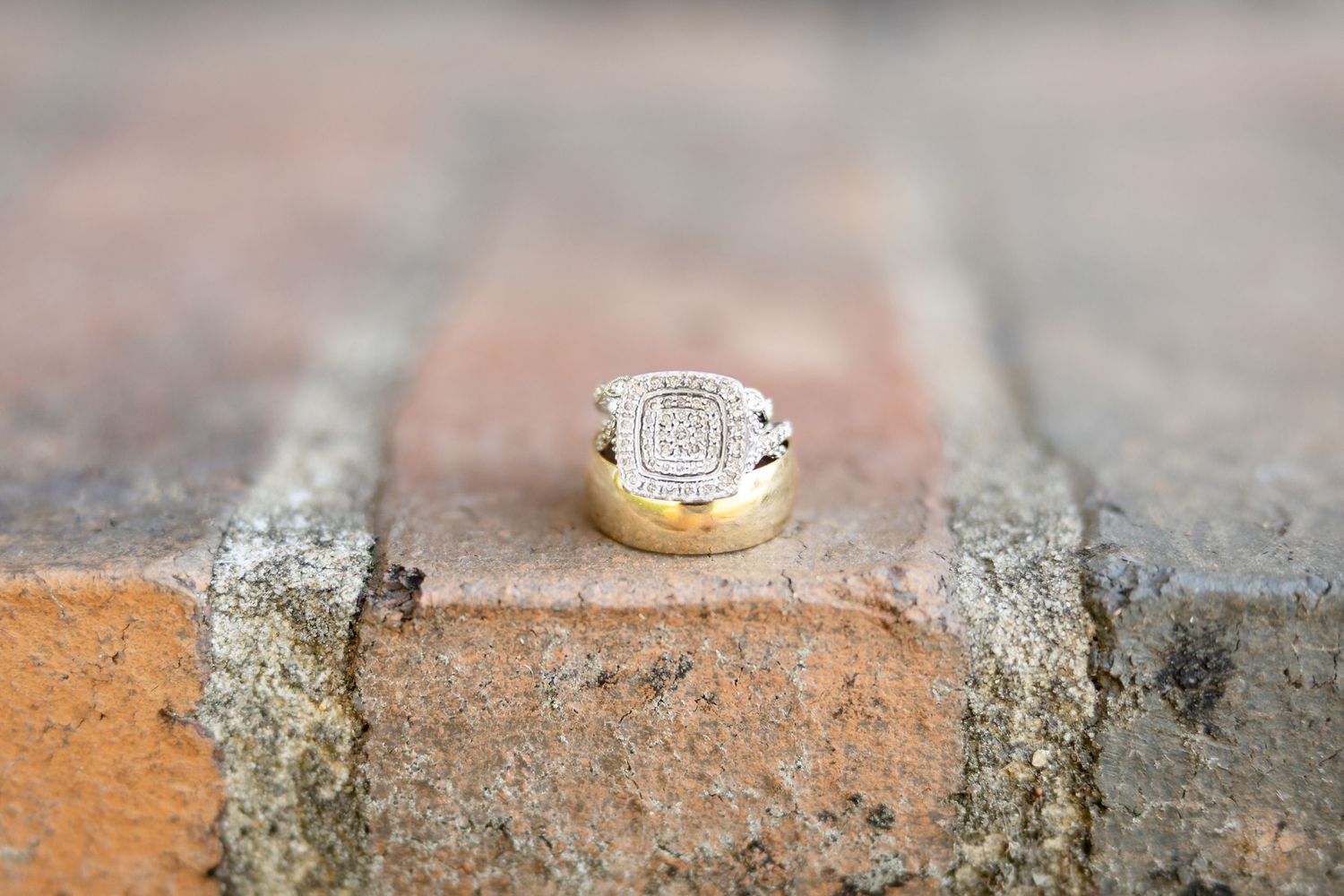 Diamond engagement ring resting on a plain gold wedding band displayed on a rustic brick surface.